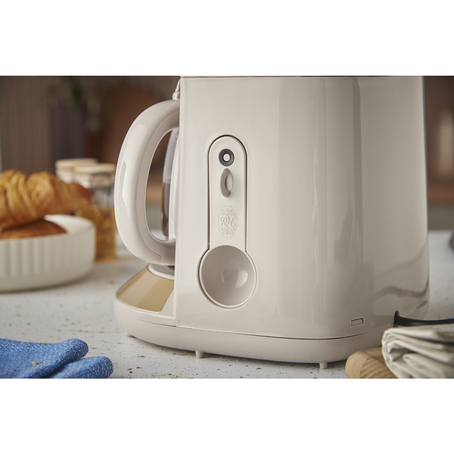 A beige coffee maker sits on a countertop. A croissant and bowl are in the background. A blue oven mitt rests on the counter.