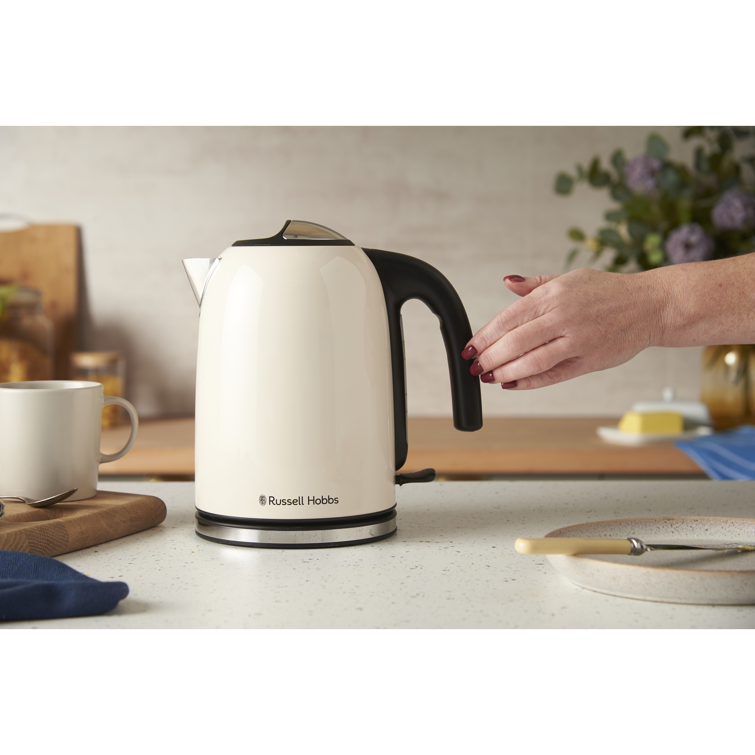 A cream-colored kettle with a black handle sits on a counter. A hand reaches for the handle. A mug, plate, and flowers are also visible.