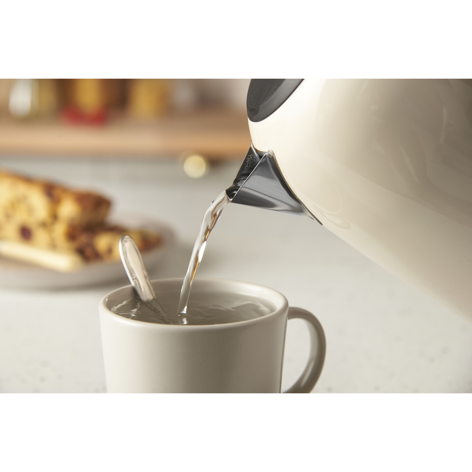 Water pouring from a cream-colored kettle into a mug with a spoon. Cake on a plate is in the background.
