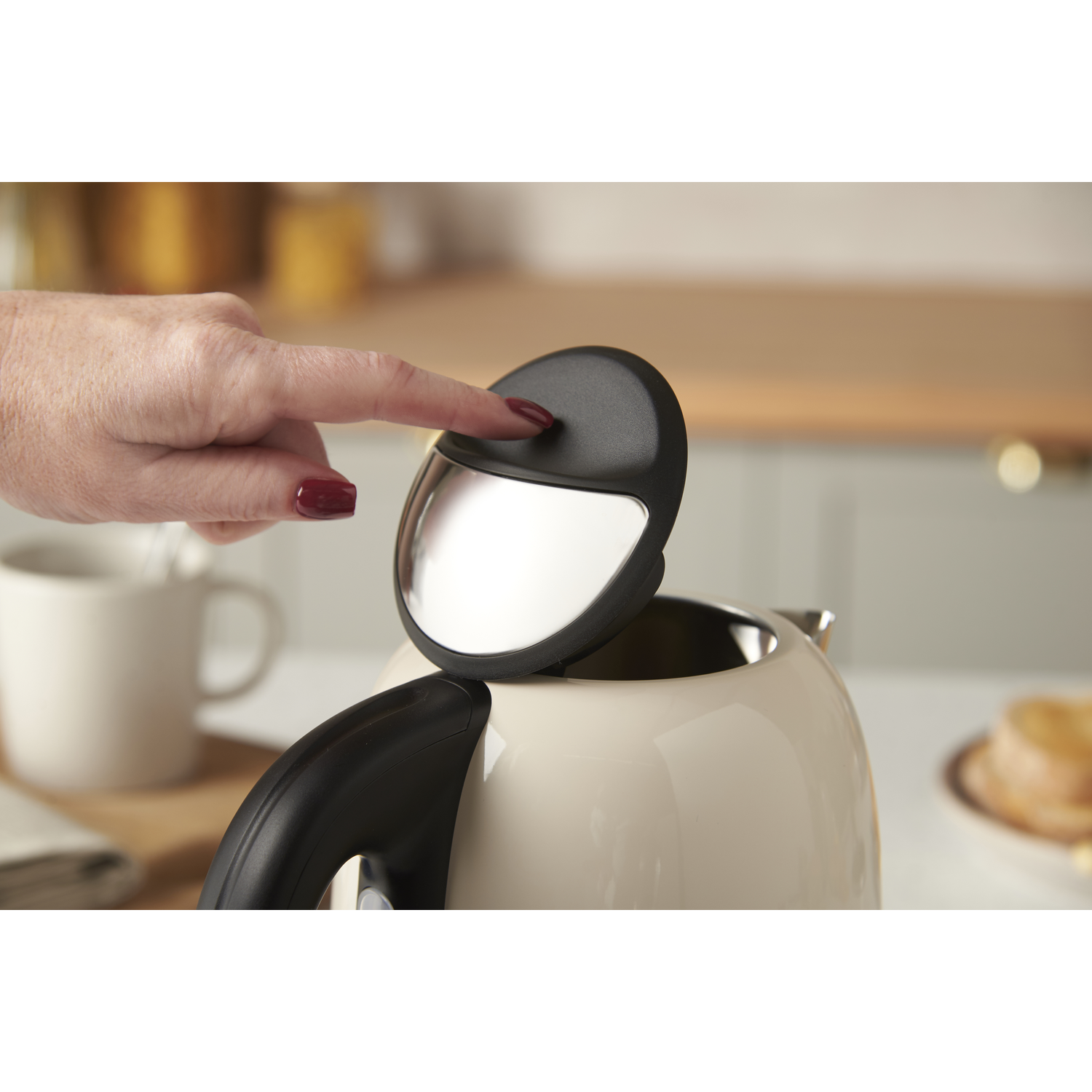 A hand presses the button of a cream-colored kettle lid. A mug and food are blurred in the background.