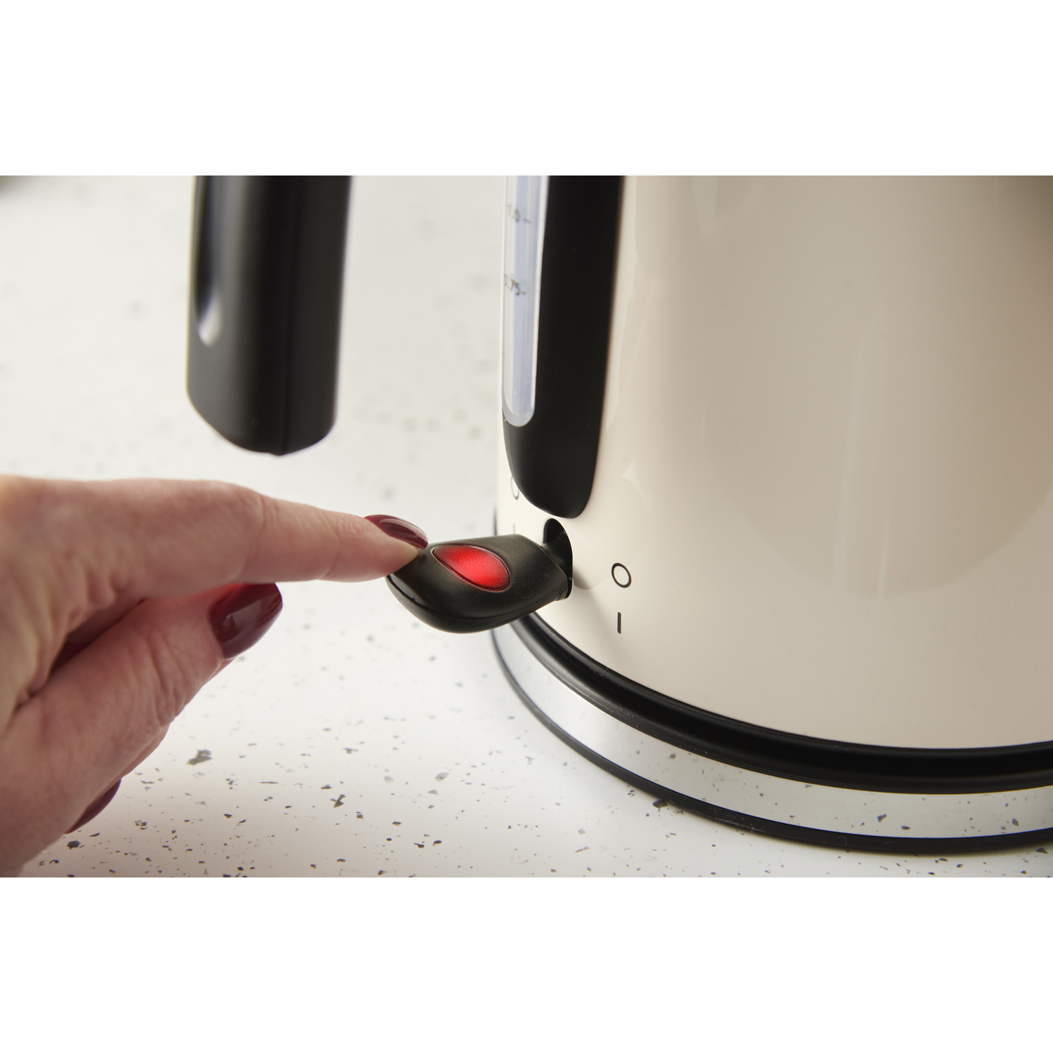 A hand presses a red-lit power button on a cream-colored electric kettle. The background has a speckled surface.