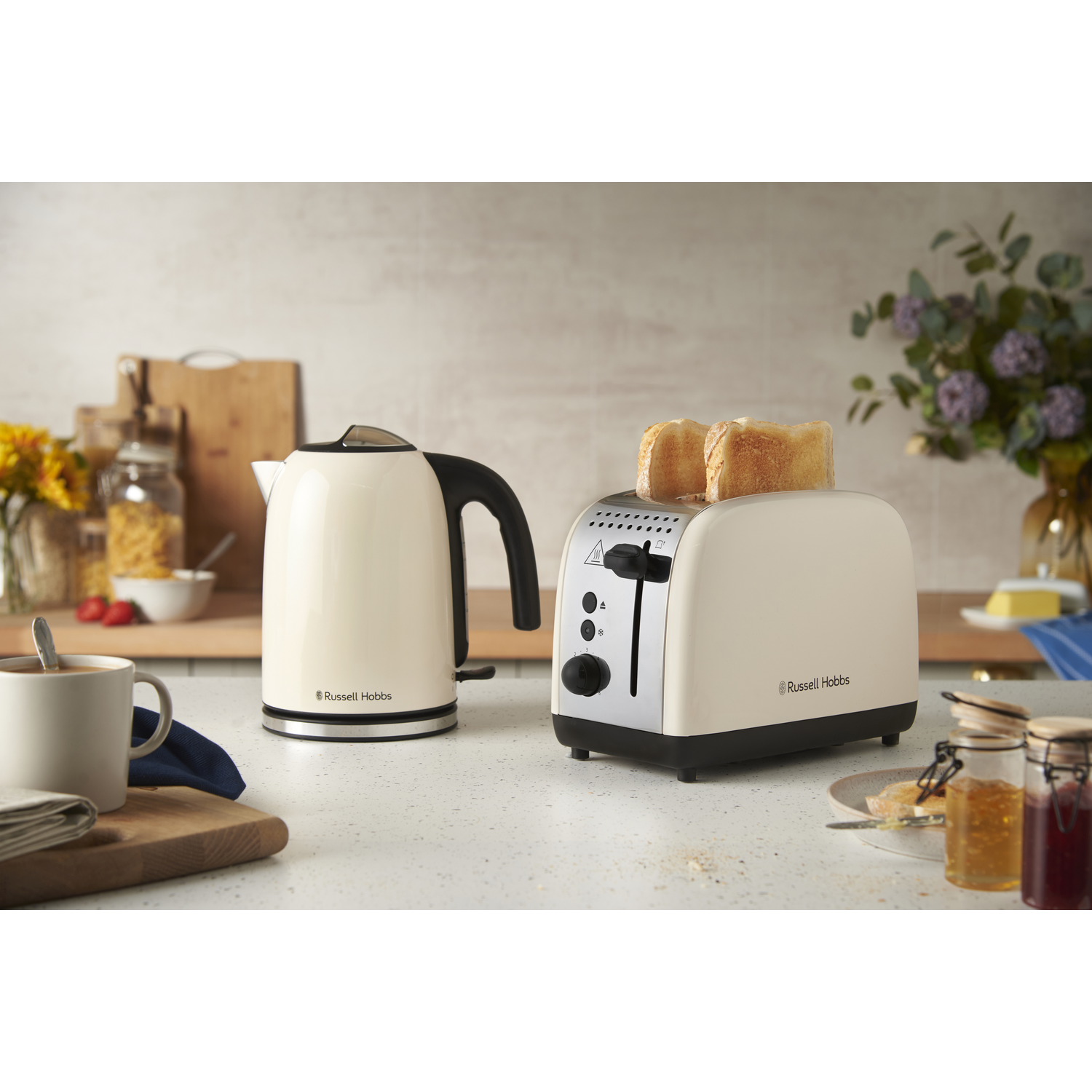 Cream-colored kettle and toaster with toast. Flowers, a mug, and jars of jam are on the countertop. A wooden cutting board is in the scene.