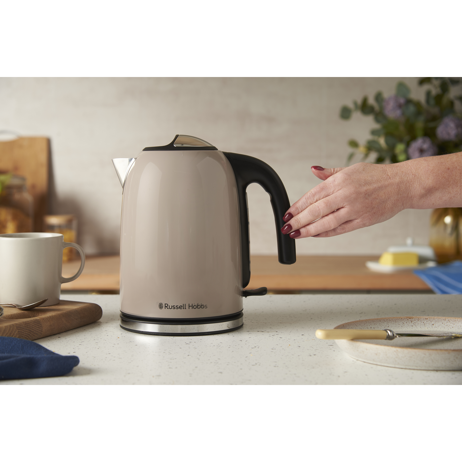 Une bouilloire beige, une main qui la tend, une tasse, une planche à découper, une assiette avec un couteau et un vase de fleurs sont sur le comptoir.
