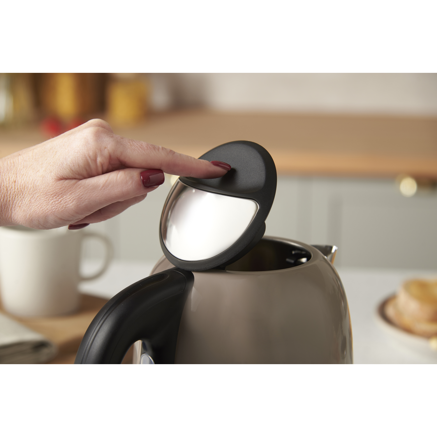Une main ouvre le couvercle d'une bouilloire. La bouilloire est grise. Une tasse et de la nourriture sont en arrière-plan.