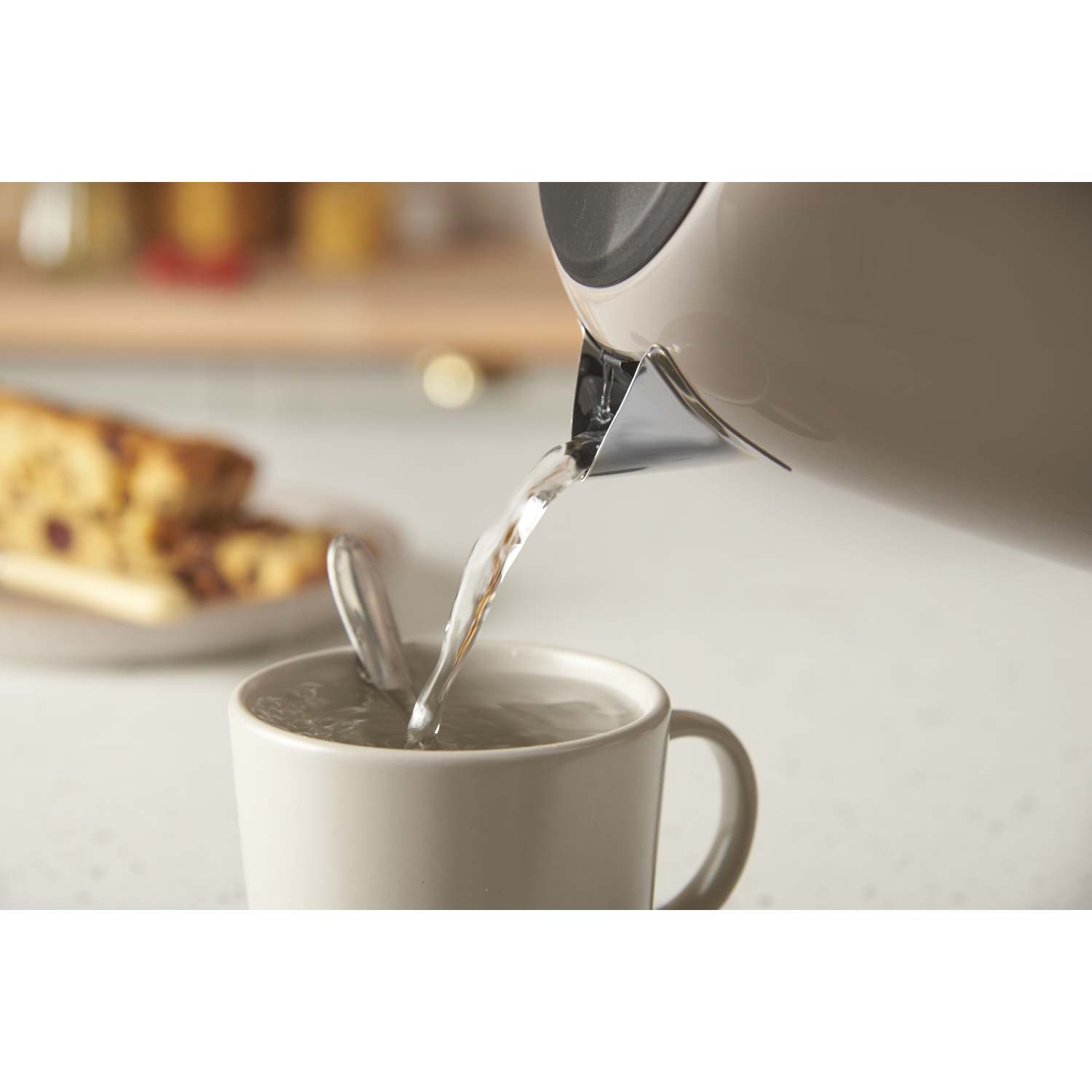 L'eau coule d'une bouilloire beige dans une tasse beige avec une cuillère. Une assiette avec un gâteau en arrière-plan.