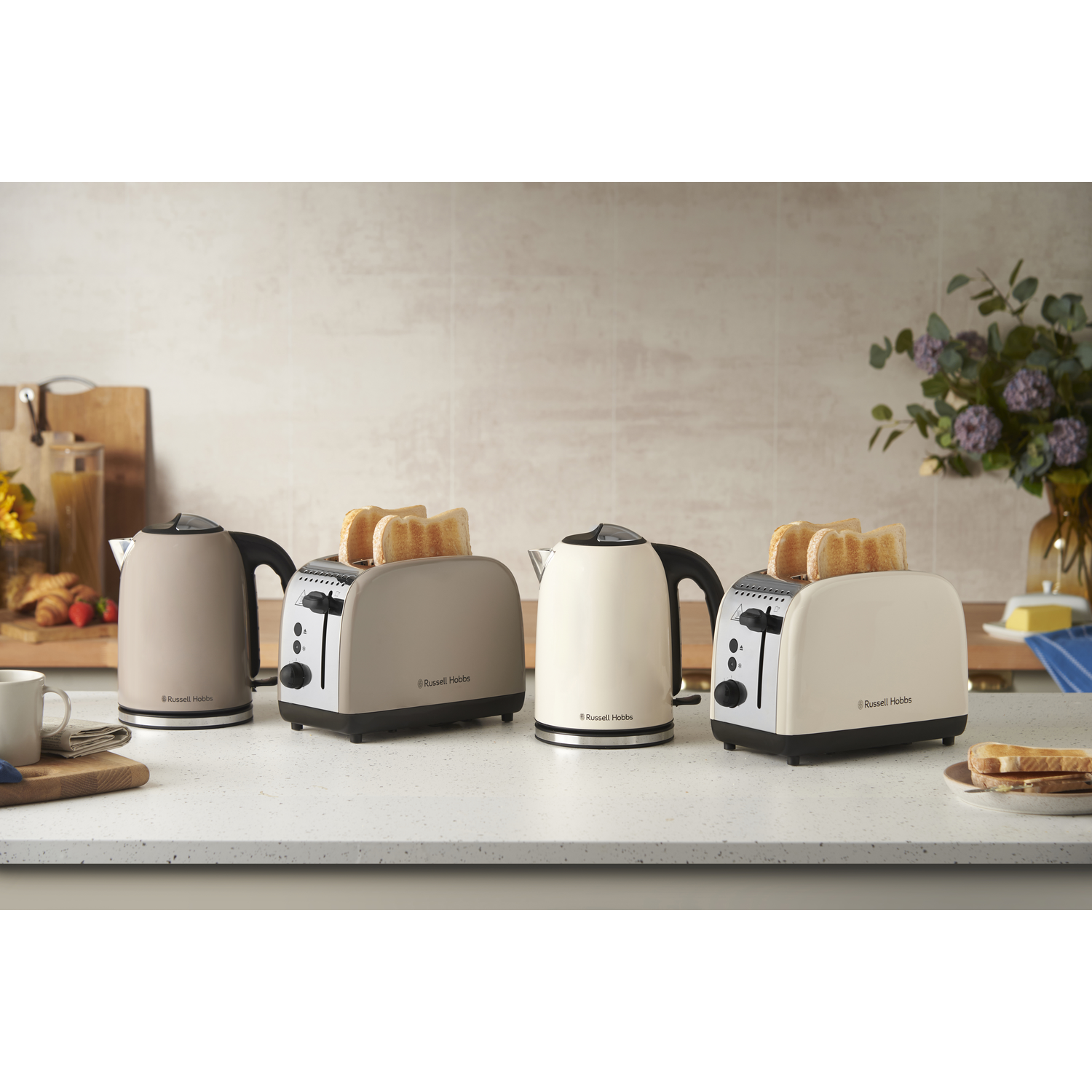 Quatre appareils Russell Hobbs : deux grille-pain avec du pain et deux bouilloires sur un comptoir, avec une composition florale en arrière-plan.