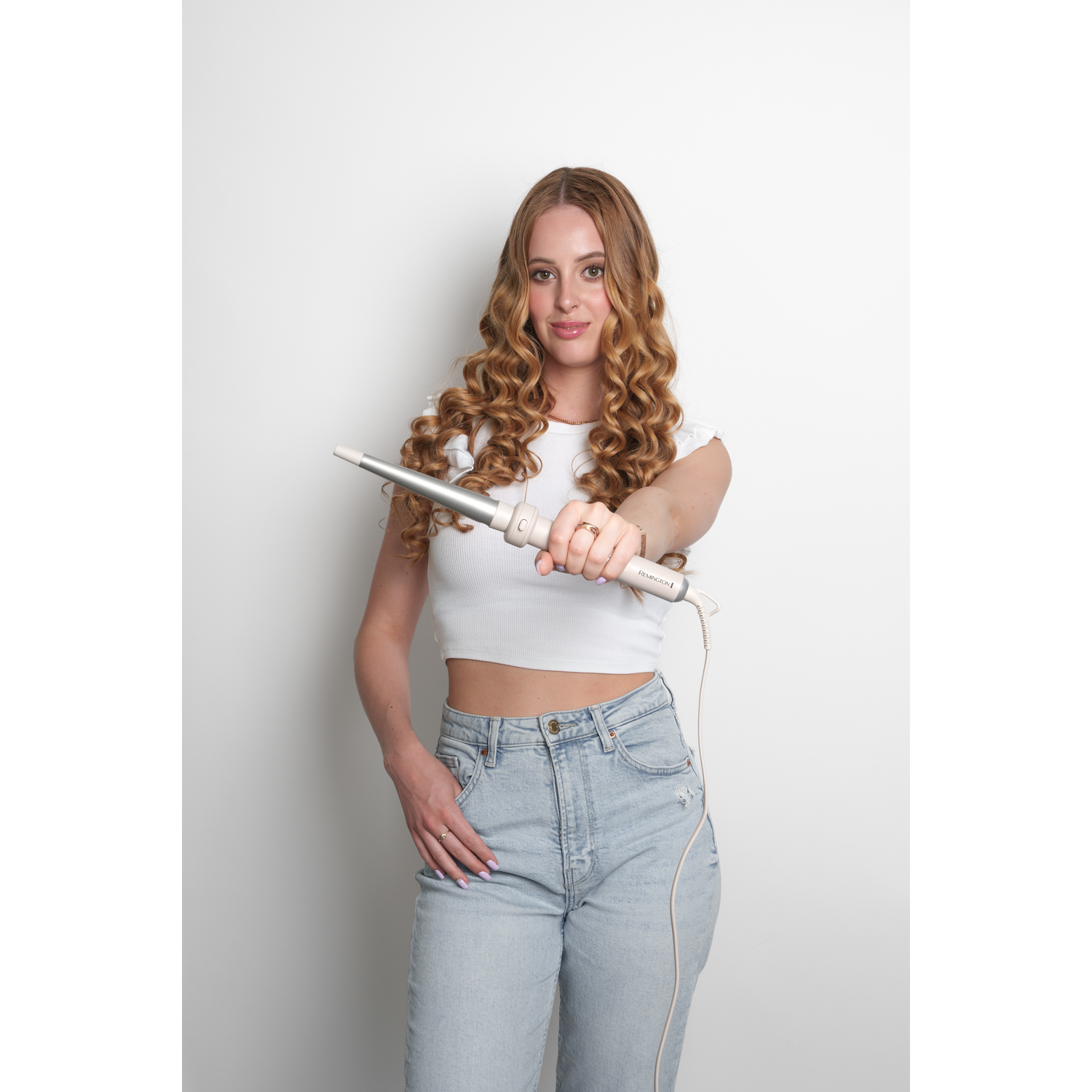 Woman with curly hair holds a curling iron. She wears a white top and jeans, standing against a white background.
