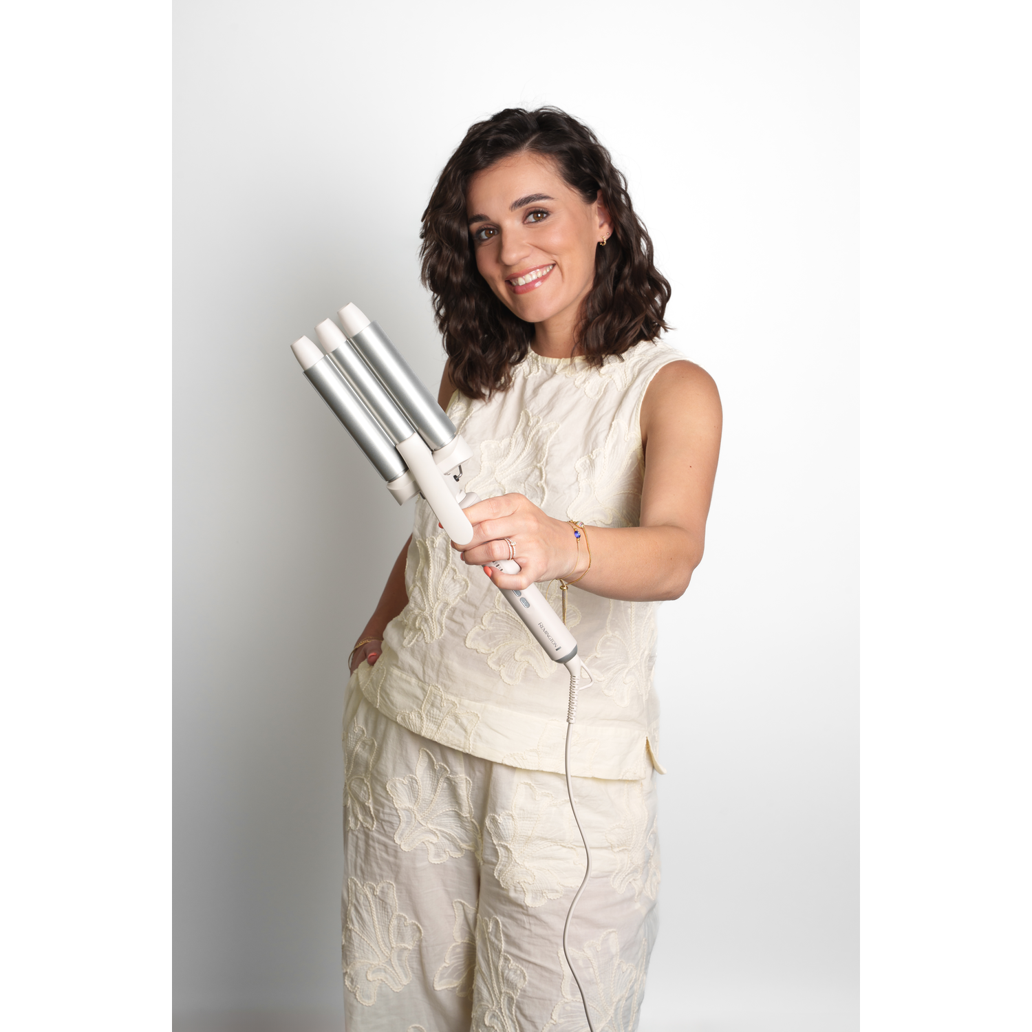 A woman holds a hair waver, smiling. She wears a cream outfit with floral embroidery. White background.