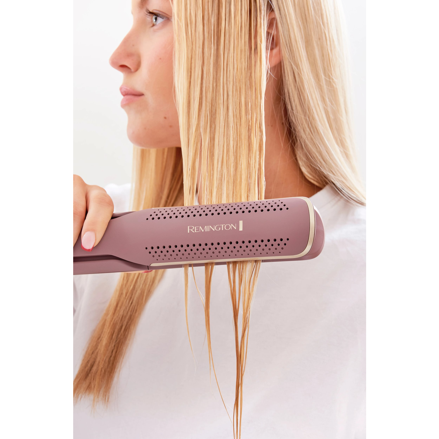 Een vrouw steilt haar blonde haar met een roze Remington stijltang. Ze draagt een wit shirt. De achtergrond is wit.