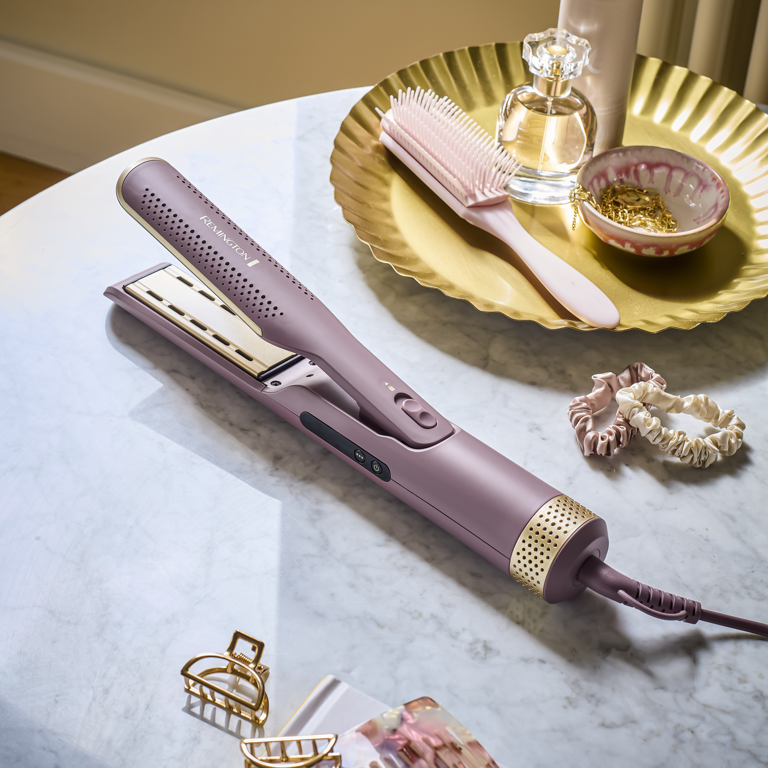 Een stijltang, haarborstel, parfum, sieraden en accessoires liggen op een marmeren tafel. Kleuren zijn mauve, goud en roze.