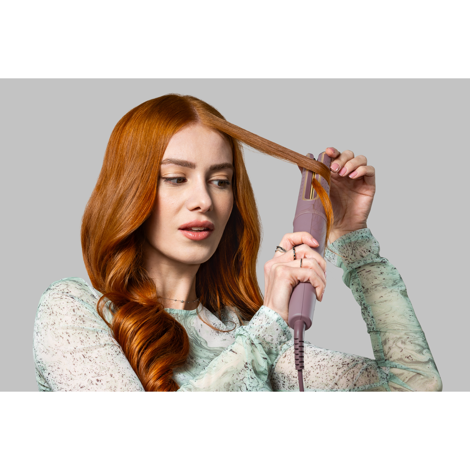 A woman with red hair uses a curling iron. She wears a patterned top and looks down. The background is a solid gray.
