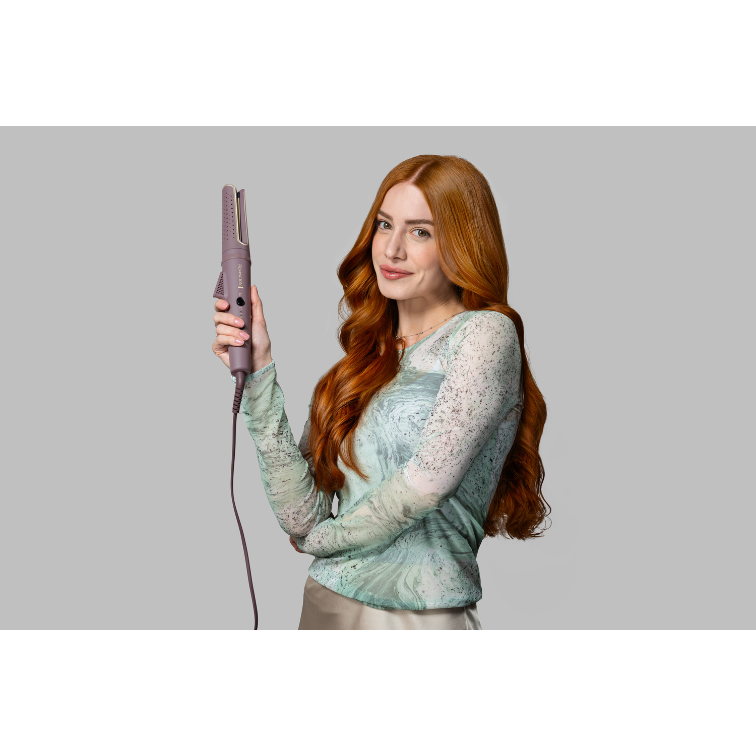 A woman with red hair holds a hair curler. She wears a patterned top. The background is grey.