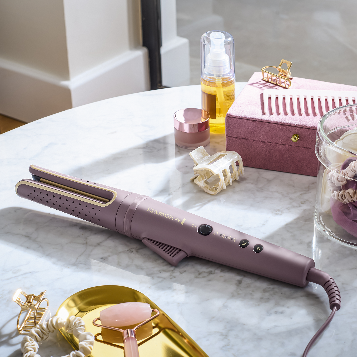 A purple Remington hair curler lies on a marble surface, next to beauty products and accessories, including a gold tray and a pink box.