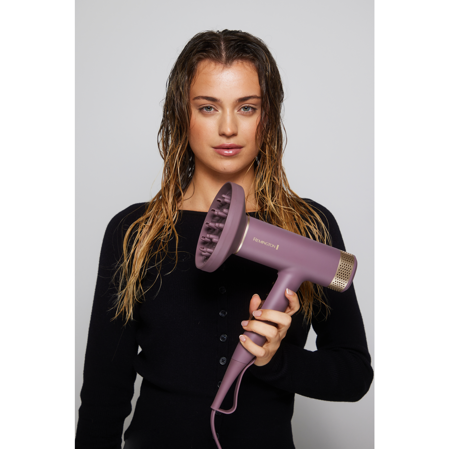 Een vrouw houdt een paarse Remington-föhn vast. Ze draagt een zwart shirt en heeft nat haar. De achtergrond is wit.
