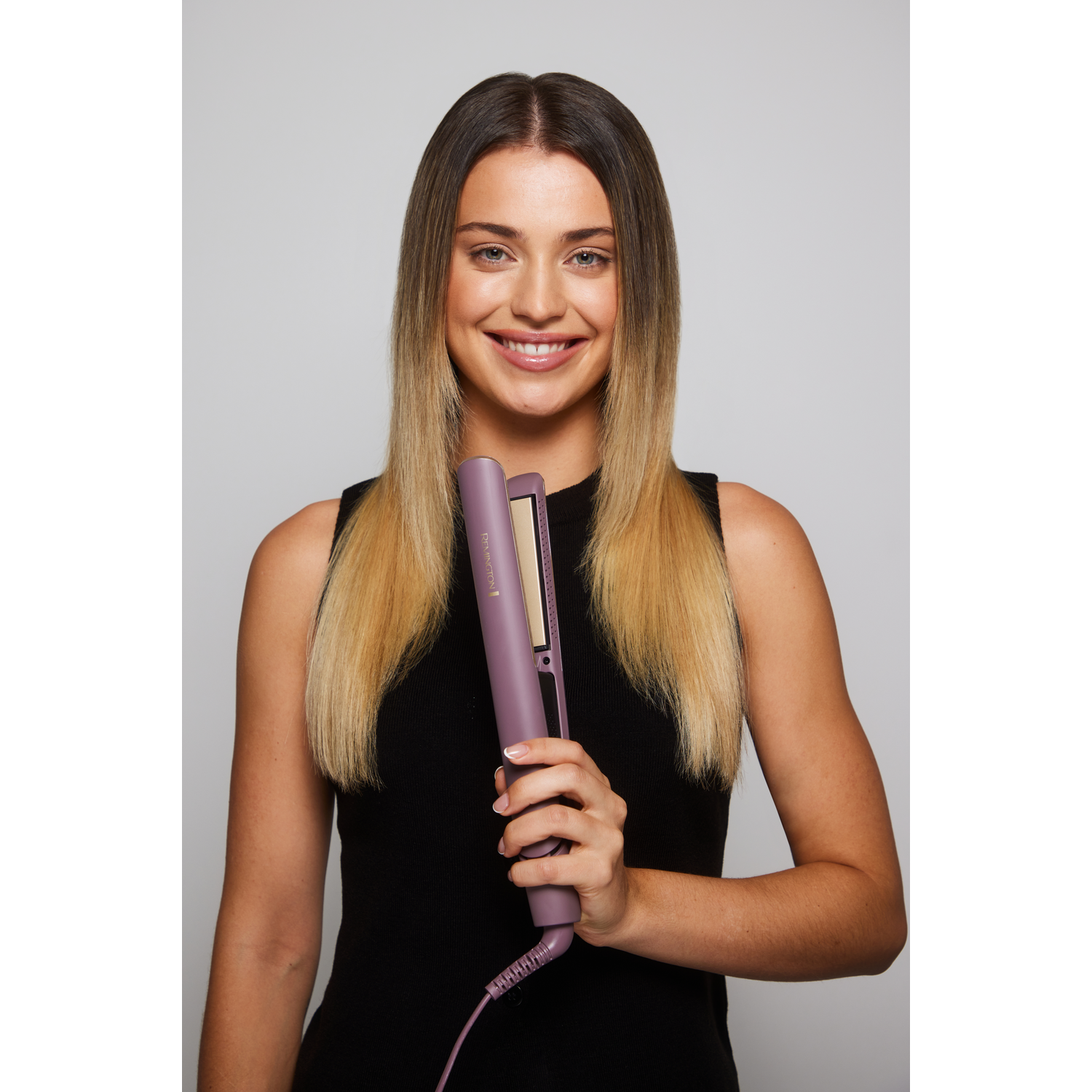 A woman holds a purple hair straightener. She wears a black top and smiles. Her blonde hair is straight. The background is plain.
