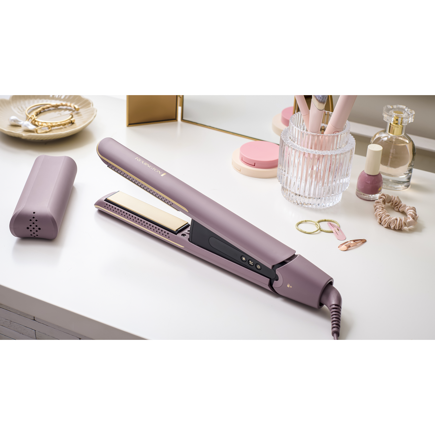 A flat iron, makeup brushes, and cosmetics are on a white table. The flat iron is open. Other accessories are visible.