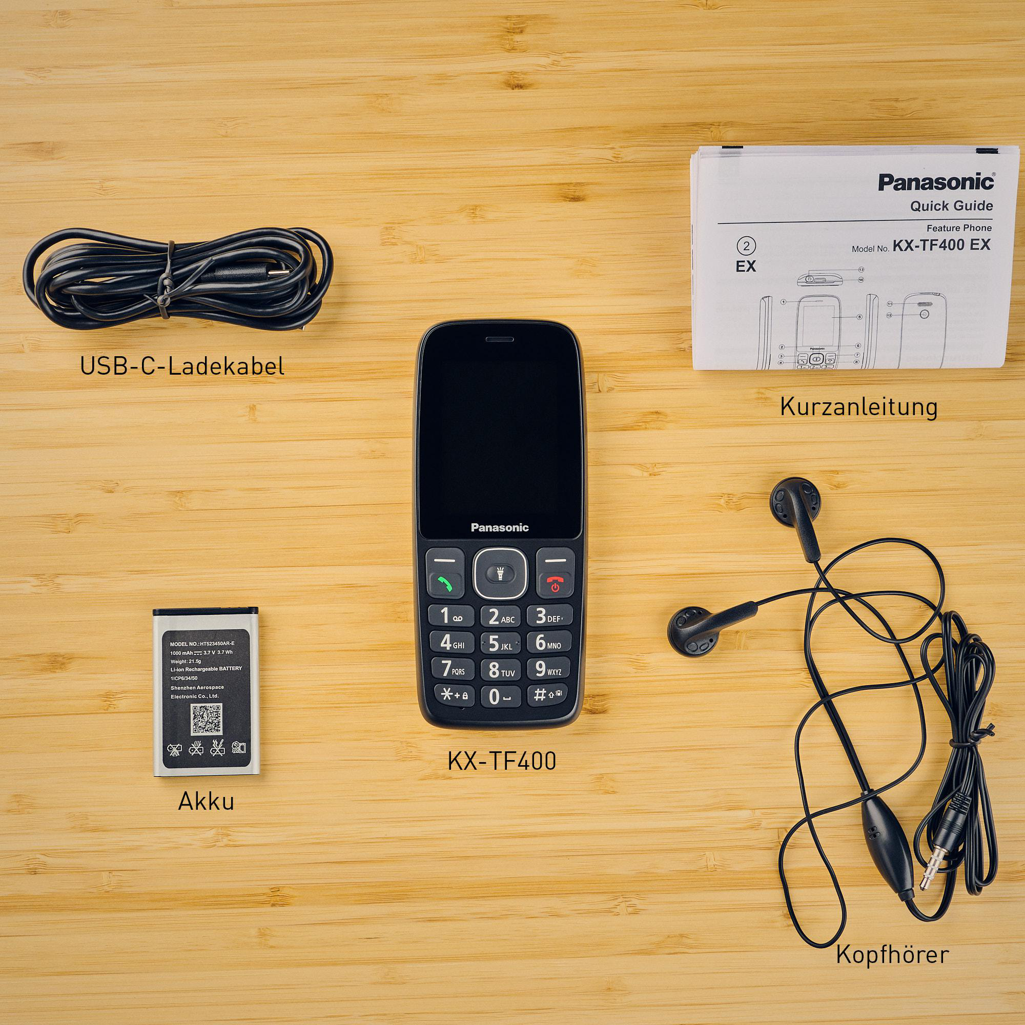 Ein Panasonic KX-TF400 Telefon mit Zubehör: Ladegerät, Akku, Kopfhörer und Handbuch auf Holzmaserung.