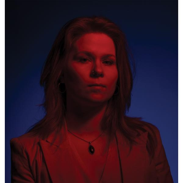 Eine Frau mit rot beleuchteter Haut, Blazer und Halskette, blickt vorwärts vor blauem Hintergrund.