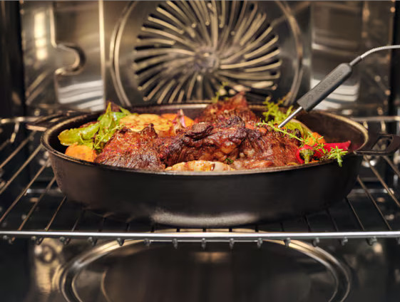 Wewnątrz piekarnika, żeliwna patelnia zawiera pieczone mięso i warzywa. Termometr do żywności jest włożony w mięso. Piekarnik jest włączony.