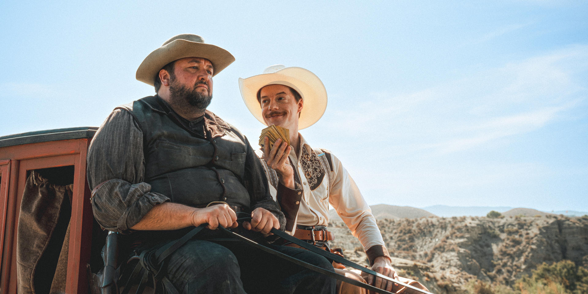 Zwei Männer mit Cowboyhüten sitzen in einer Kutsche, einer hält Karten. Eine Wüstenlandschaft im Hintergrund unter blauem Himmel.
