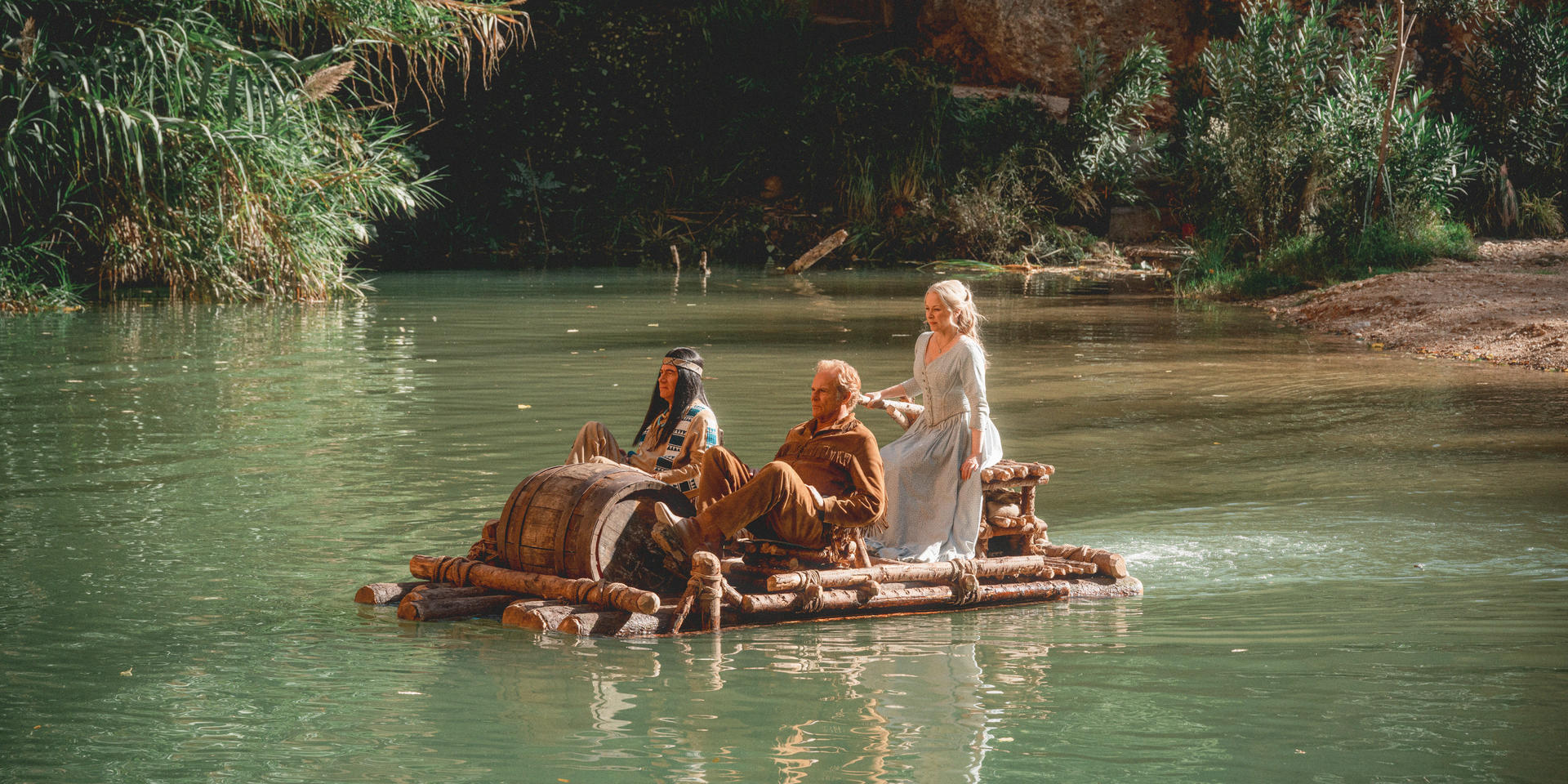 Drei Personen auf einem Floß in einem Fluss. Einer trägt indianische Kleidung, einer ein braunes Hemd und einer ein blaues Kleid. Üppige grüne Ufer umgeben.