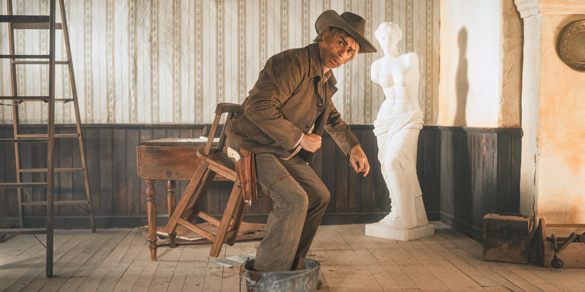 Ein Cowboy in brauner Jacke und Hut steht in einem Metalleimer. Eine Statue und ein Holztisch sind ebenfalls vorhanden.