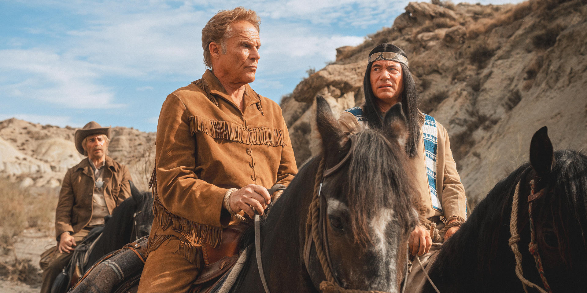 Drei Männer auf Pferden in einer Wüstenlandschaft. Einer trägt eine Jacke mit Fransen, ein anderer einen Indianerkopfschmuck. Ein dritter Mann trägt einen Cowboyhut.