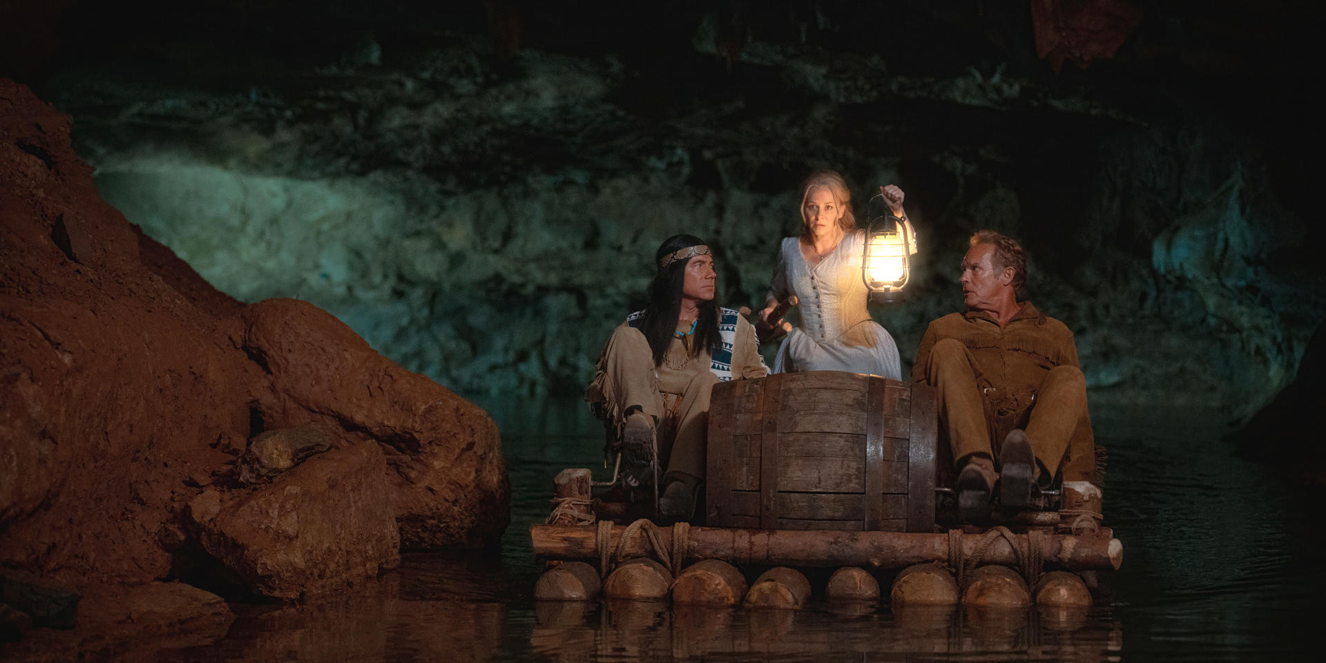 Drei Personen auf einem Floß in einer Höhle. Einer hält eine Laterne. Dunkle Höhlenwände und Wasser. Mann mit Stirnband.