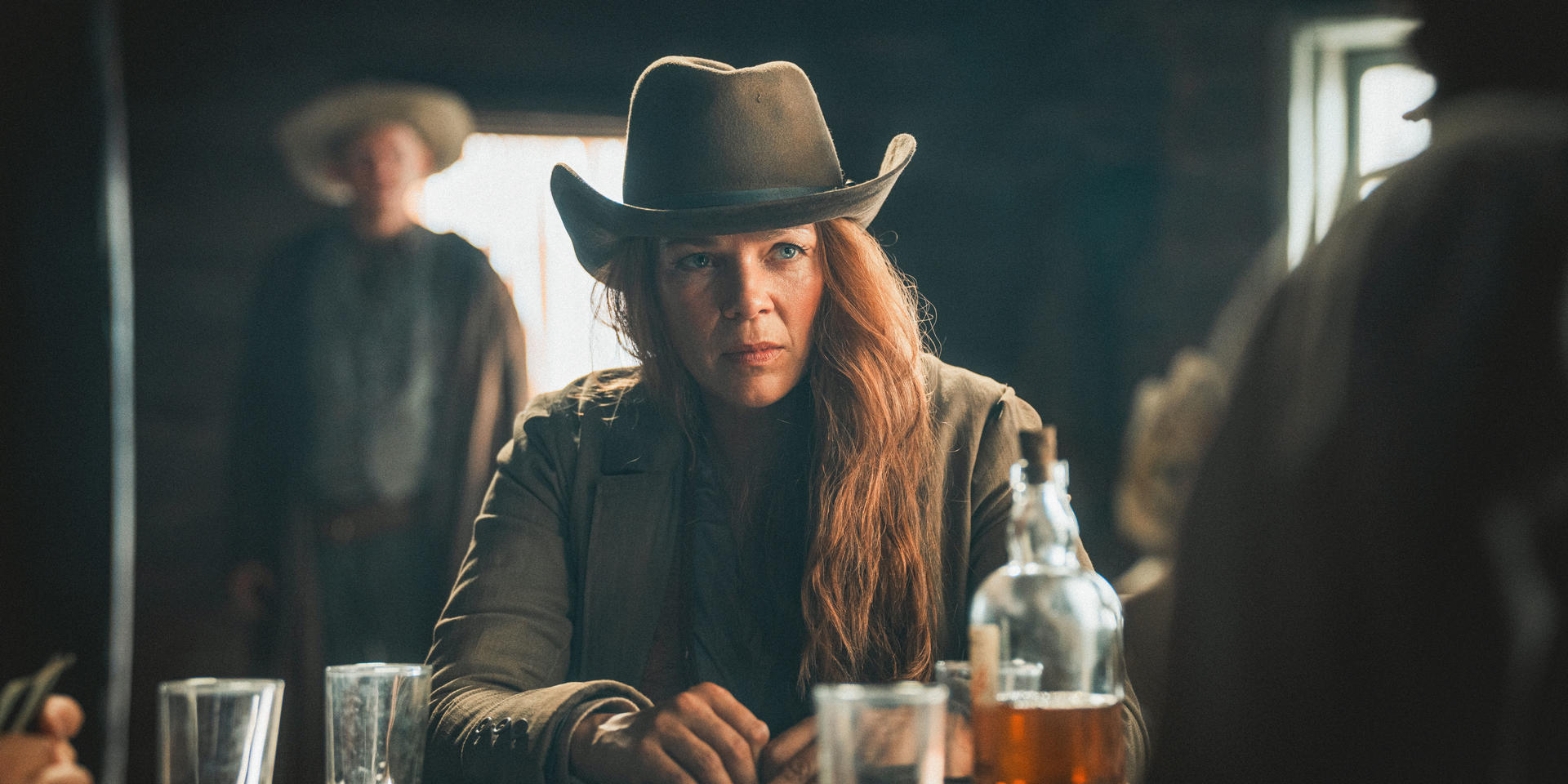 Eine Frau mit Cowboyhut sitzt an einem Tisch und schaut aufmerksam. Eine Flasche und Gläser stehen vor ihr. Andere sind im Hintergrund.