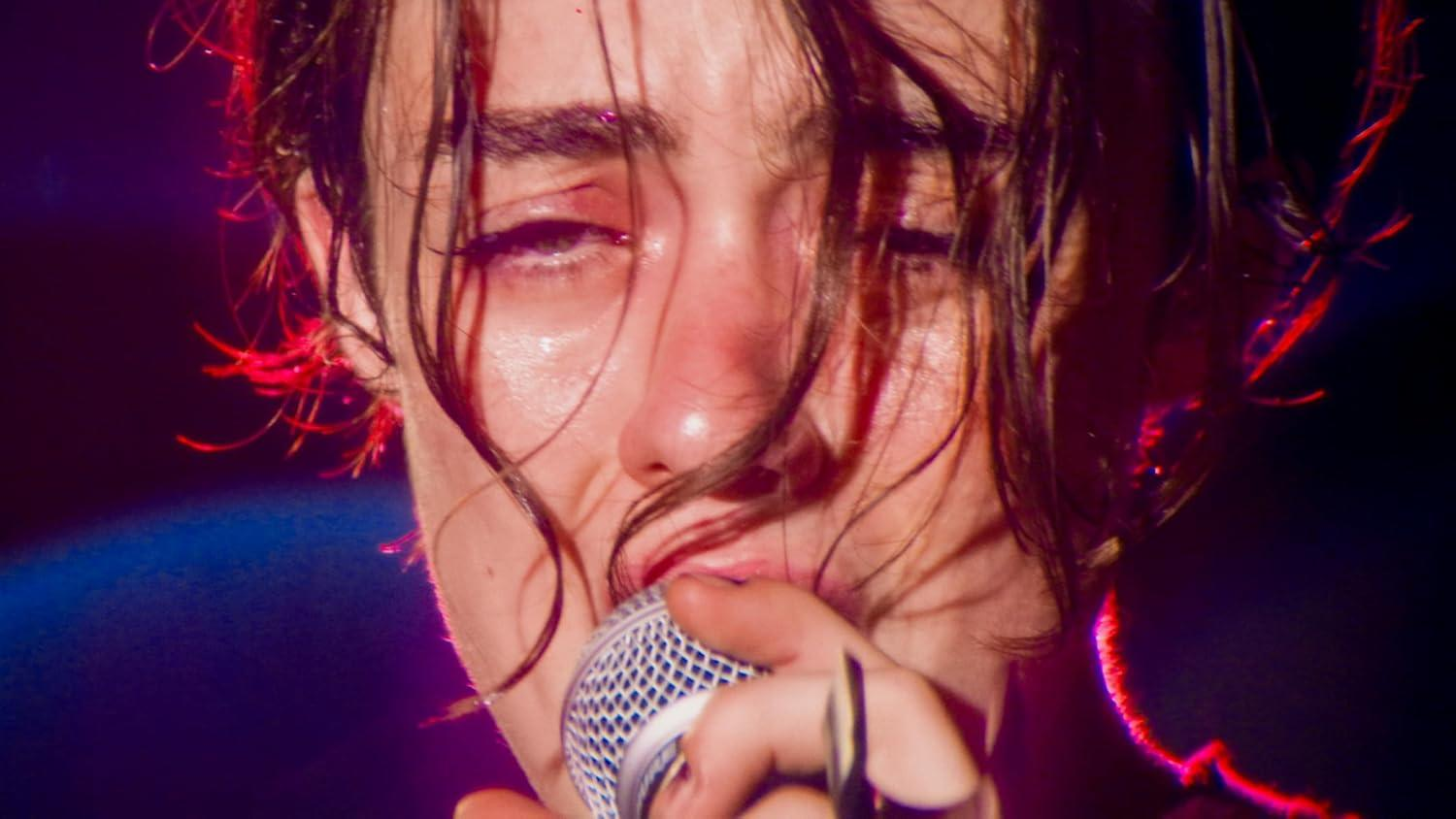 Nahaufnahme einer Person, die in ein Mikrofon singt. Gesicht ist beleuchtet, mit nassem Haar. Rote Bühnenlichter sind sichtbar.