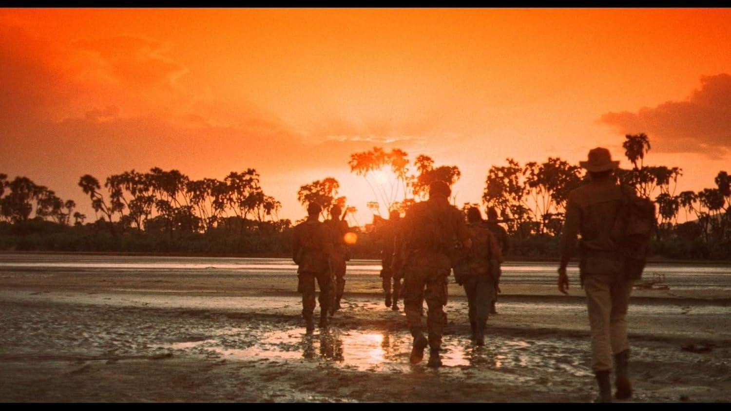 Eine Gruppe von Soldaten geht am Sonnenuntergang an einem nassen Strand entlang. Silhouetten vor orangefarbenem Himmel und Bäumen im Hintergrund.