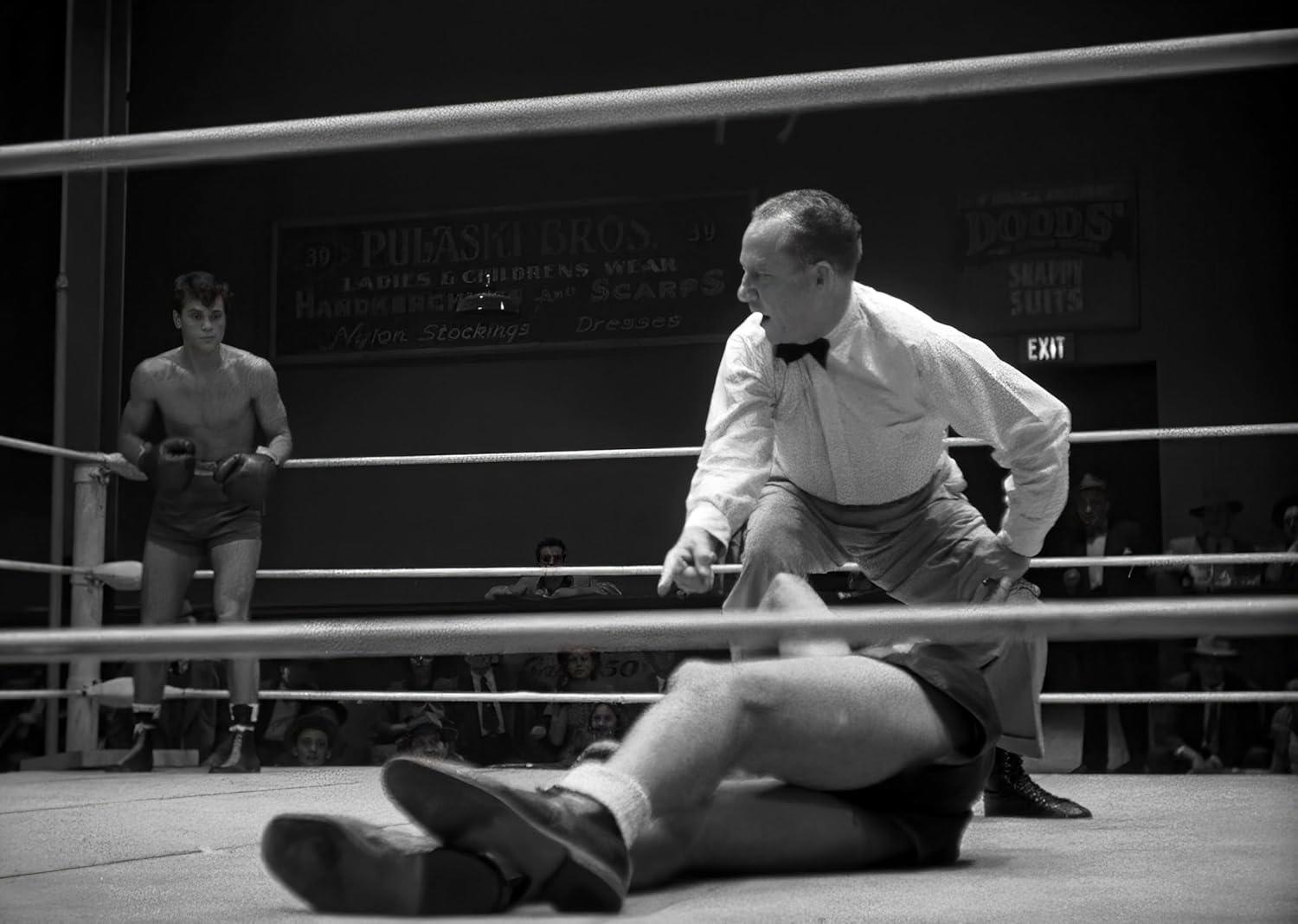 Boxer am Boden, Schiedsrichter beugt sich über ihn. Ein anderer Boxer steht im Ring. Schilder an der Wand: Pulaski Bros, Doods und Exit.