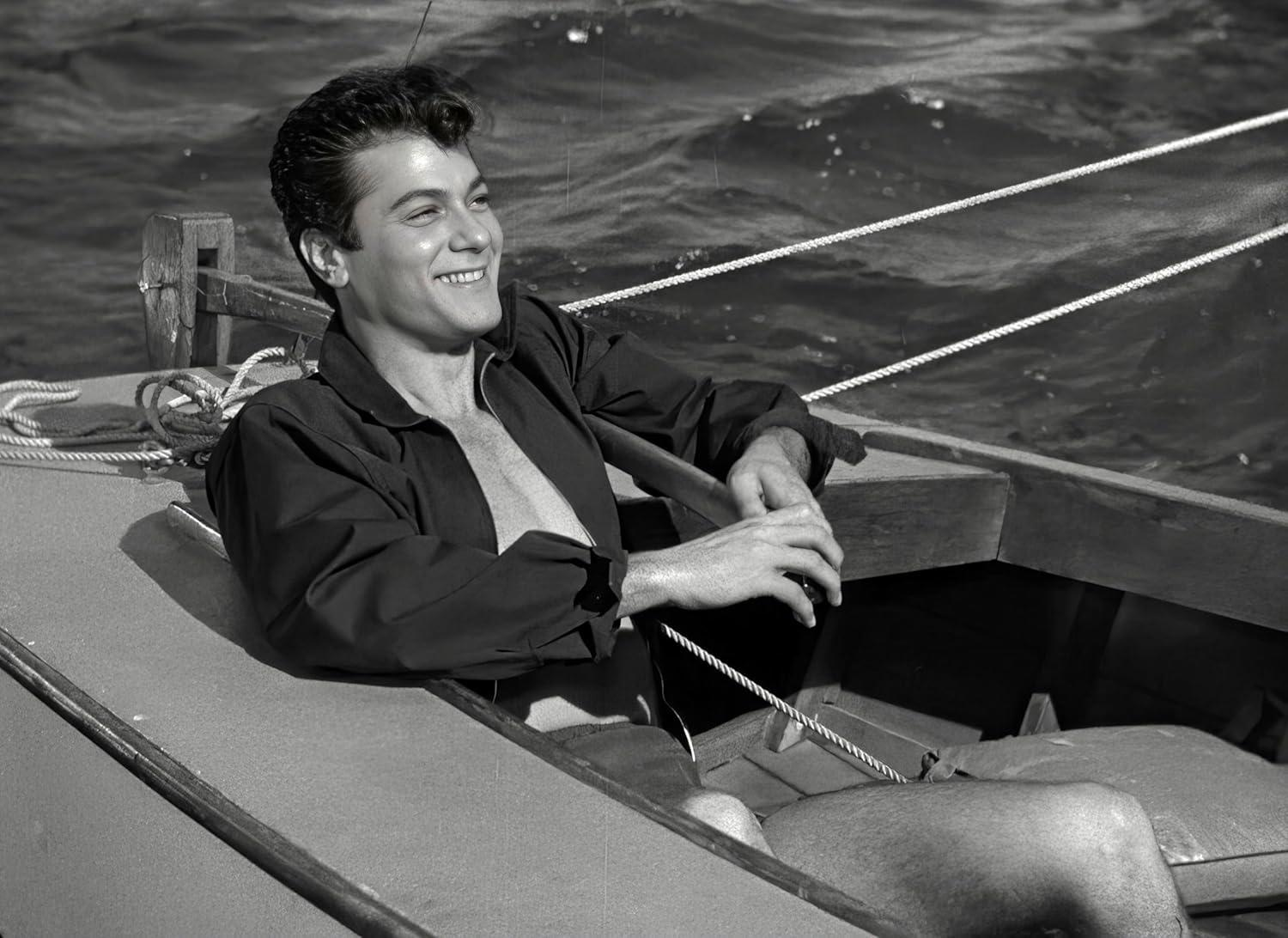 Ein Mann in einem Boot lächelt. Er trägt eine dunkle Jacke, die am Oberkörper offen ist. Der Hintergrund ist das Meer.