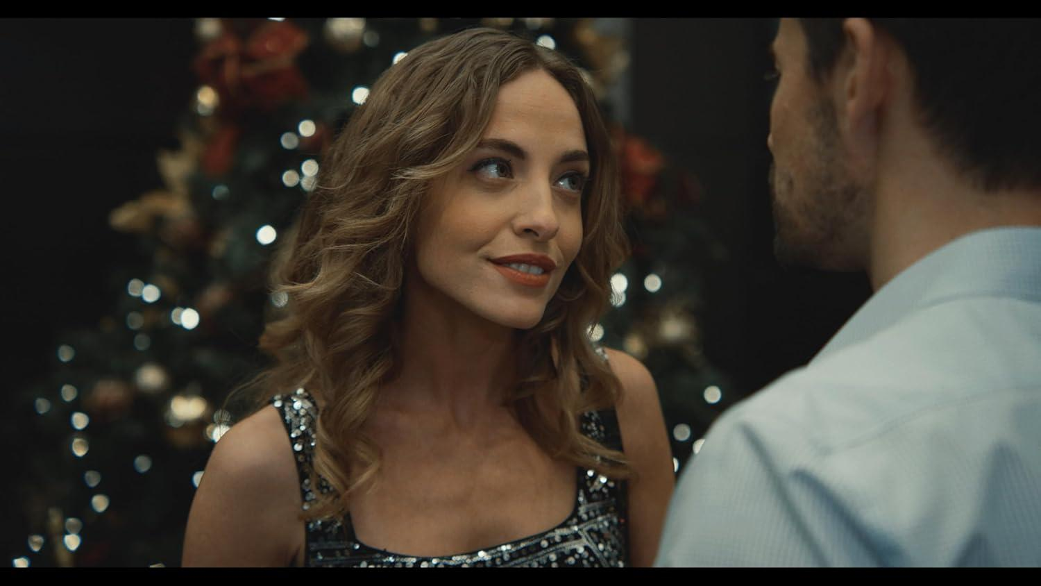 Eine Frau lächelt einen Mann in festlicher Atmosphäre an. Sie trägt ein paillettenbesetztes Oberteil, er ein blaues Hemd. Weihnachtsbeleuchtung im Hintergrund.
