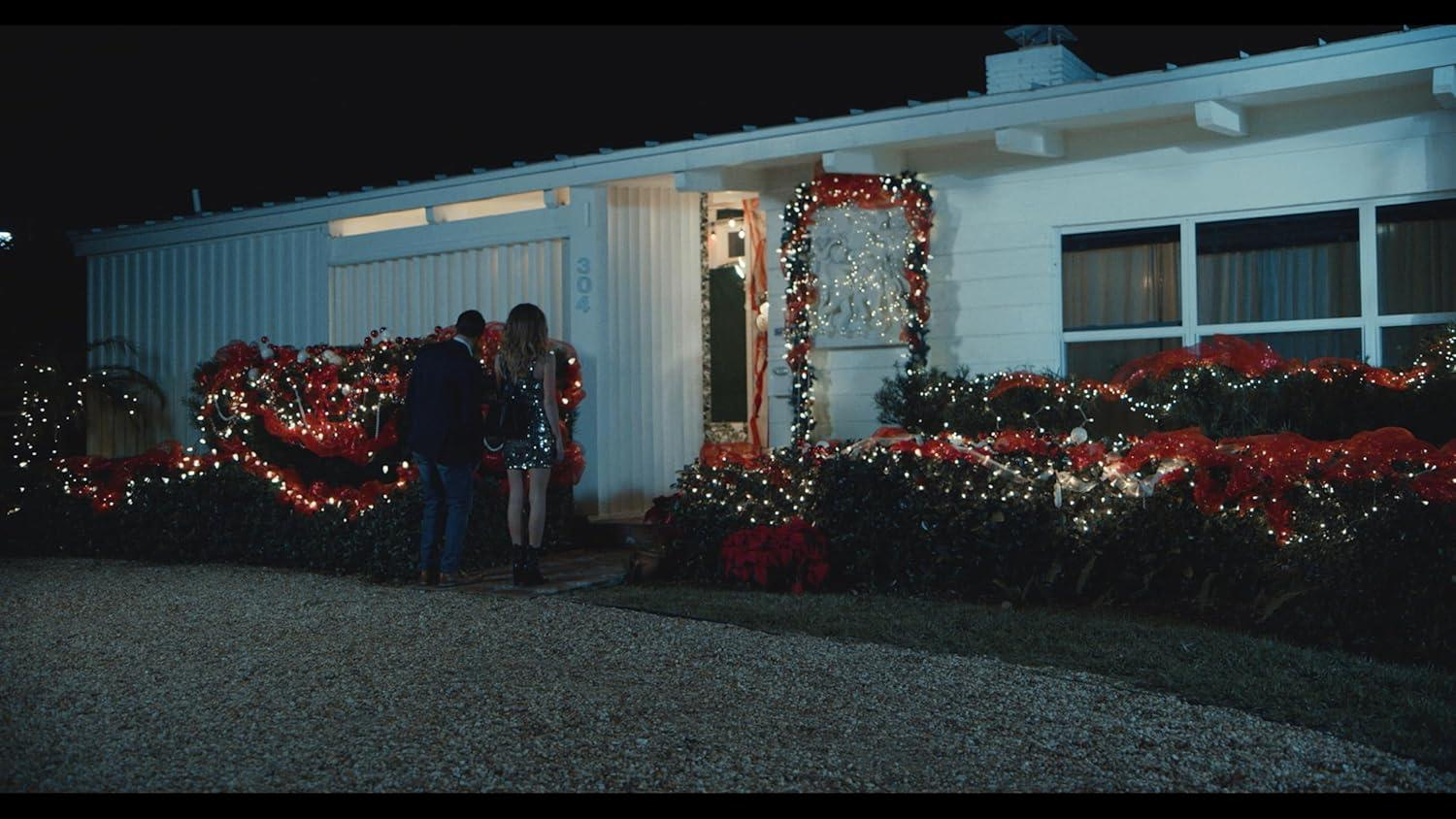 Ein Paar steht vor einem mit Weihnachtsbeleuchtung geschmückten Haus. Das Haus hat eine weiße Fassade, und der Hof hat Kies.