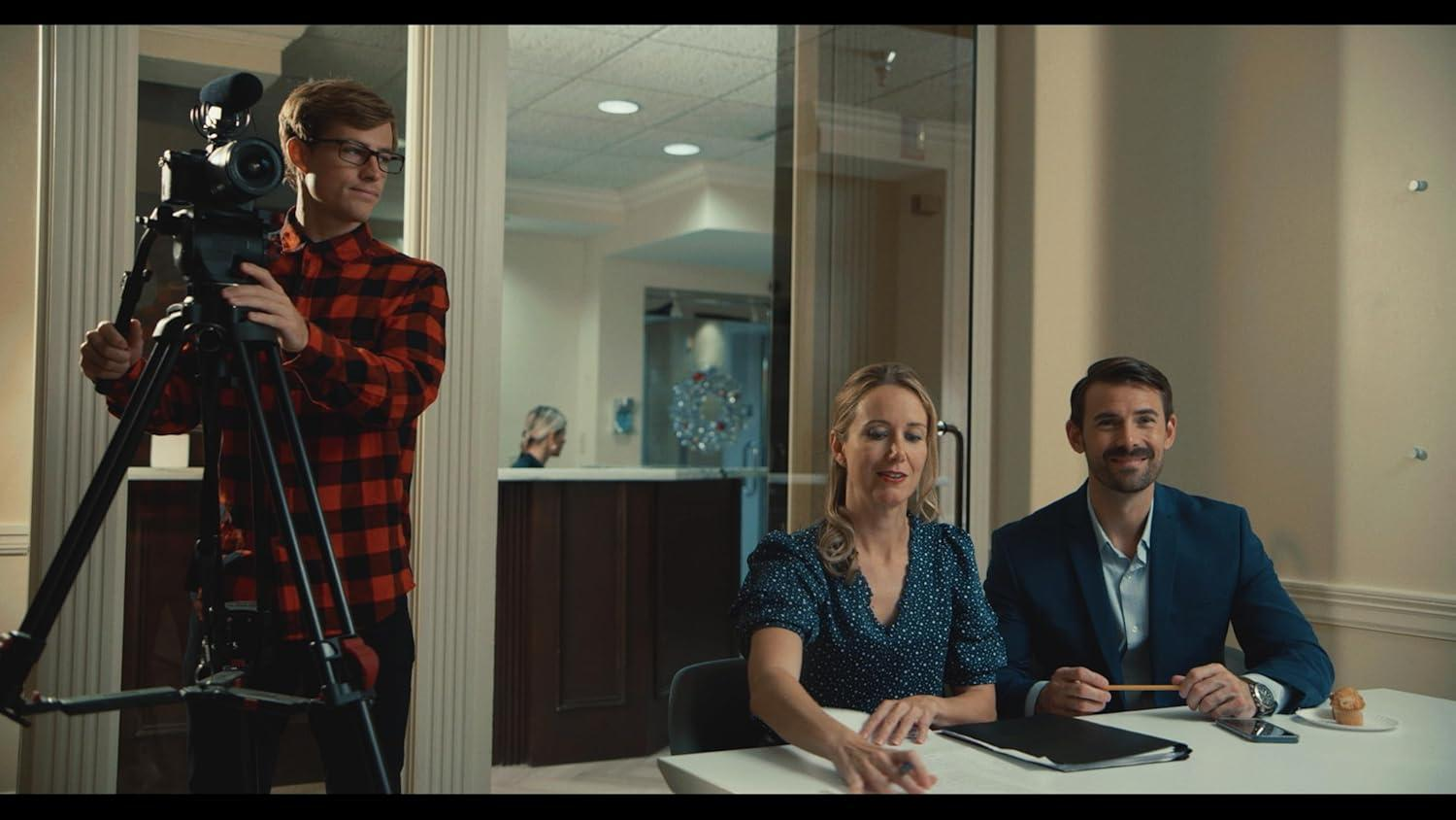 Ein Mann mit Kamera auf Stativ filmt Frau und Mann am Tisch. Im Hintergrund eine Frau.