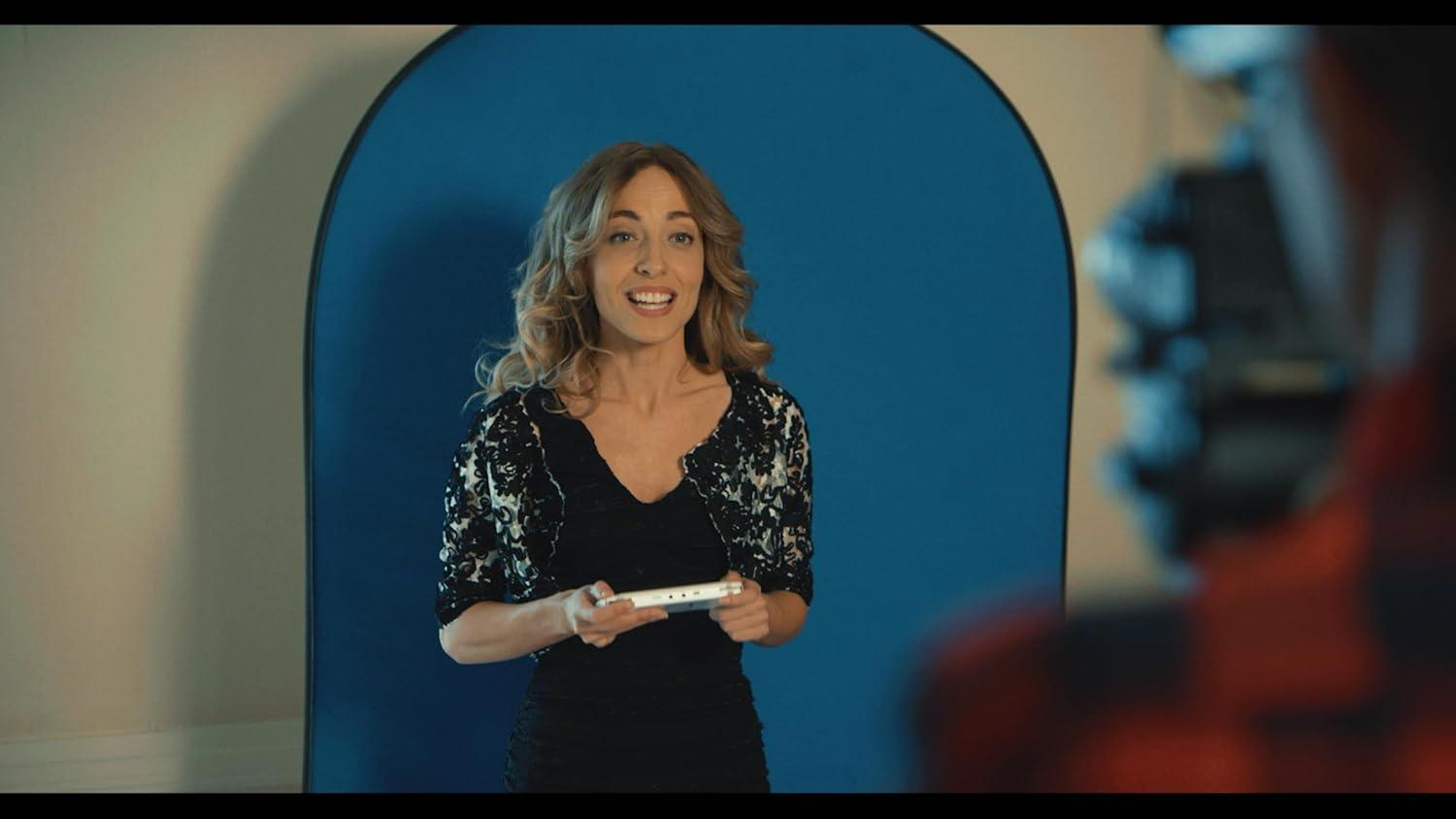 Eine Frau mit blonden Haaren, trägt ein schwarzes Kleid und eine Spitzenjacke, hält ein weißes Gerät vor einem blauen Hintergrund.
