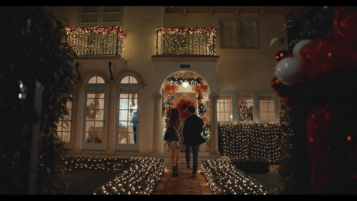 Ein Paar geht nachts auf ein dekoriertes Haus zu. Das Haus hat Weihnachtsbeleuchtung und Dekorationen. Der Vordergrund hat weitere Lichter.