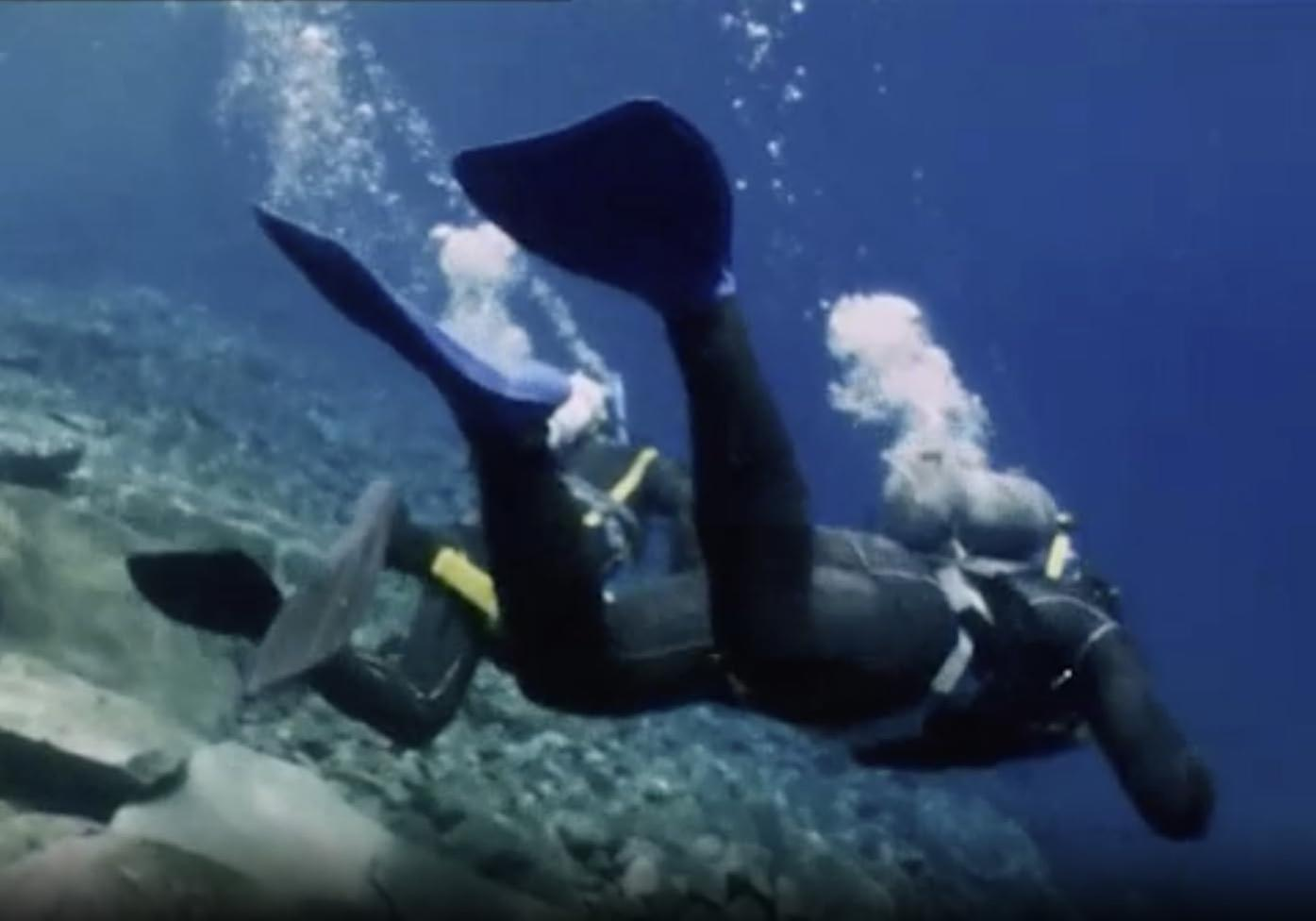 Taucher schwimmen unter Wasser, tragen schwarze Anzüge und blaue Flossen. Blasen steigen auf. Felsiger Meeresboden im Hintergrund. Das Wasser ist blau.