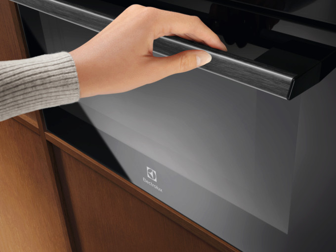 Ręka otwiera czarne drzwi piekarnika. Piekarnik ma uchwyt z metalowym wykończeniem. Widoczne jest logo Electrolux.