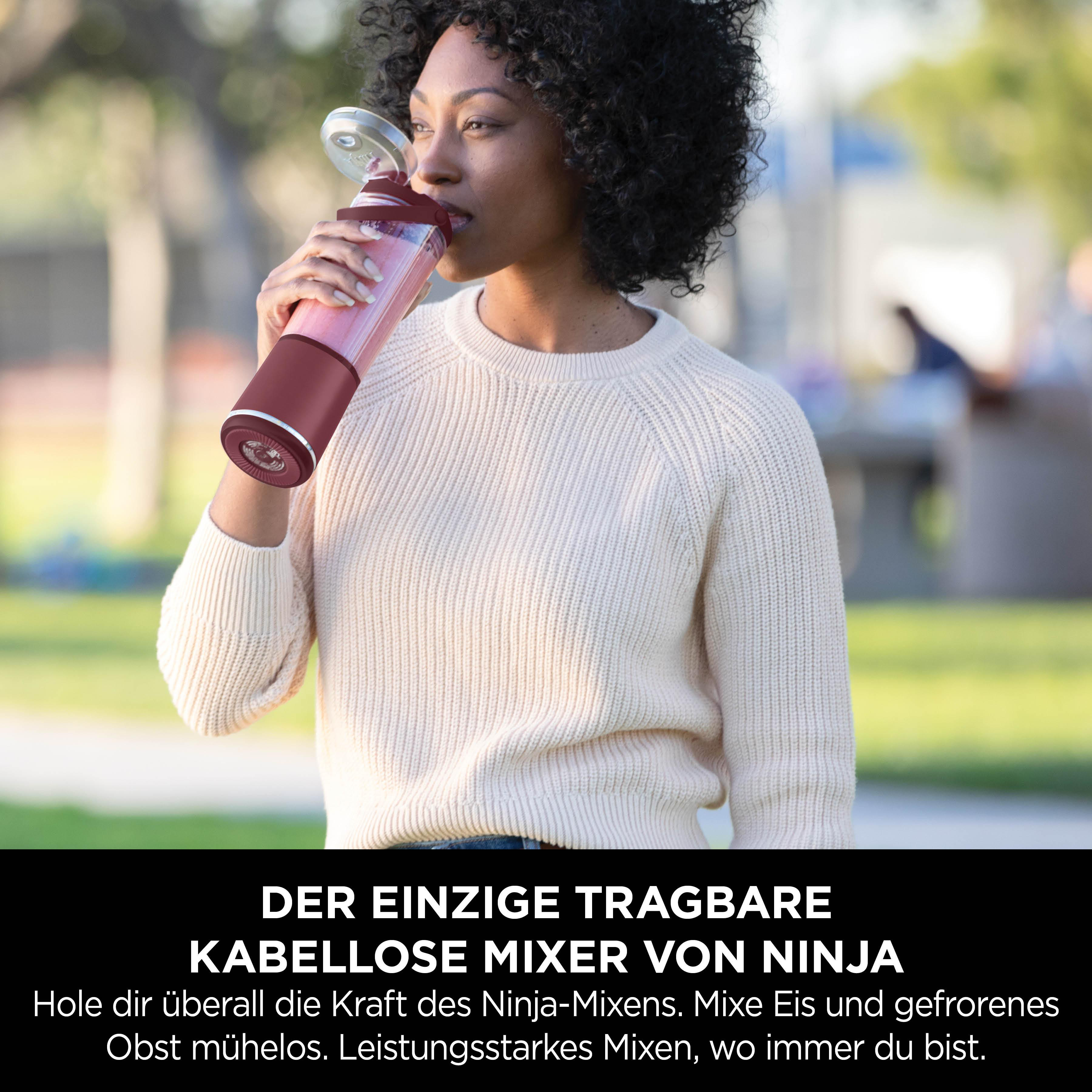 Eine Frau trinkt aus einem bordeauxroten und transparenten Mixer. Sie trägt einen cremefarbenen Pullover. Der Hintergrund ist verschwommen grün.