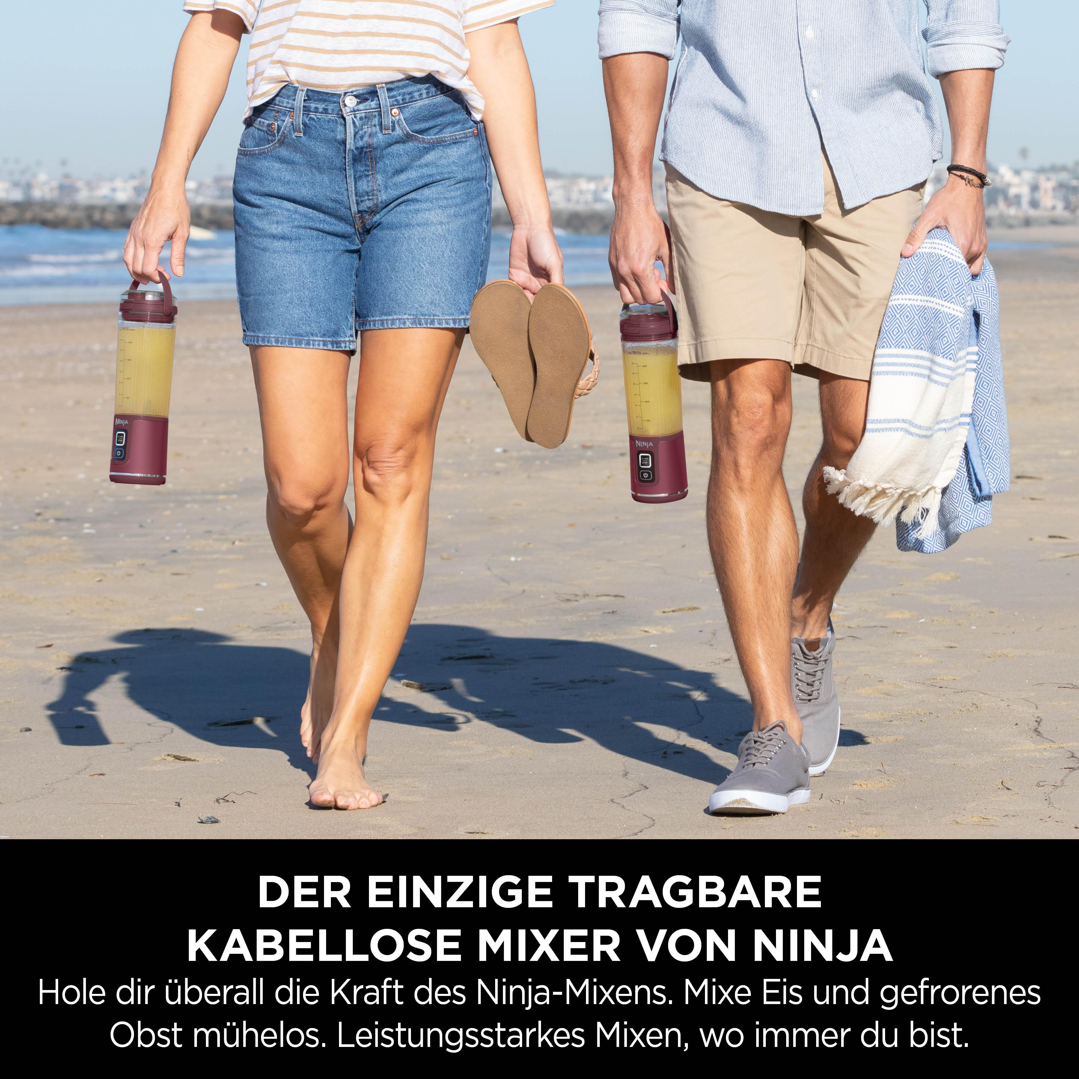 Zwei Personen gehen am Strand entlang, jede hält einen Mixer mit gelber Flüssigkeit. Einer trägt Sandalen, der andere ein Handtuch.