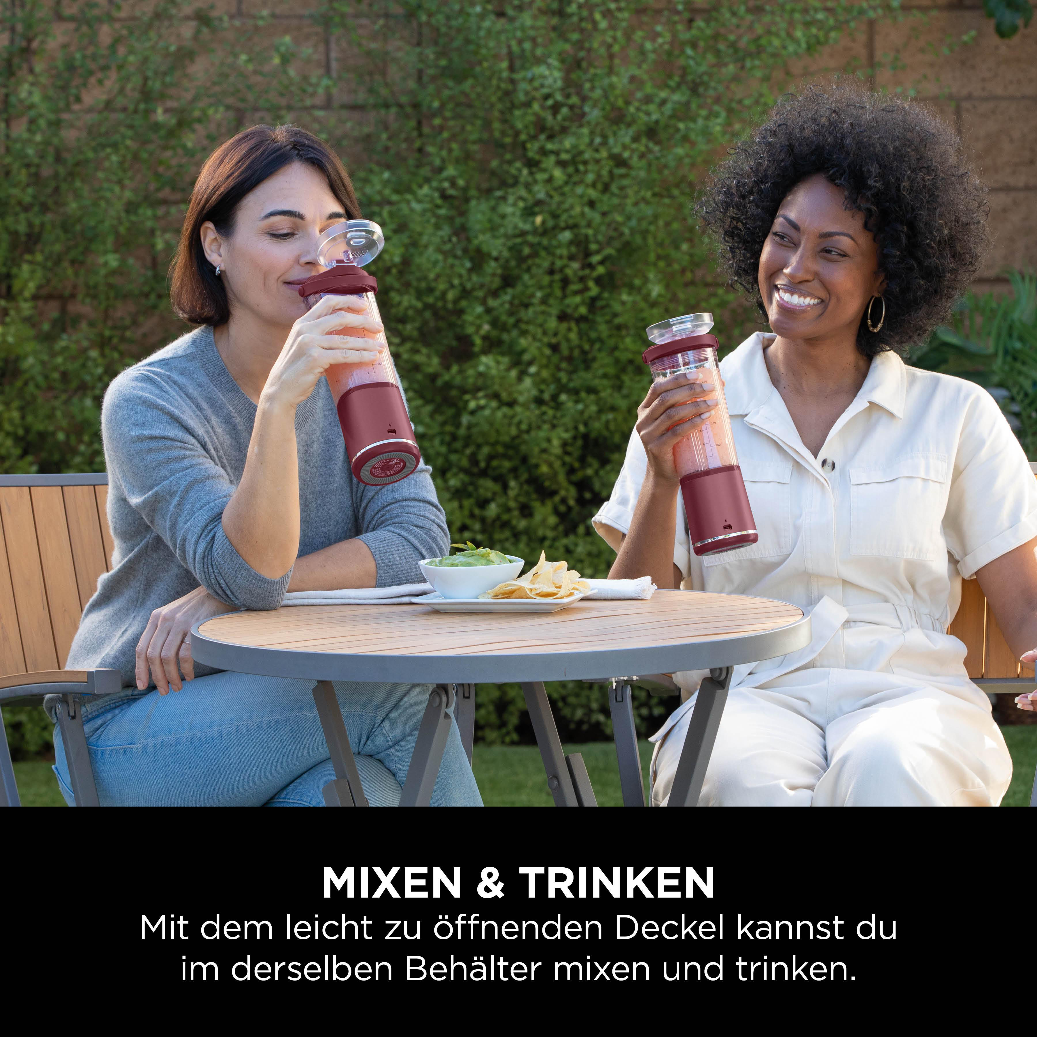 Zwei Frauen sitzen draußen und trinken aus Mixern. Auf einem Tisch stehen Guacamole und Chips. Hintergrund: grüne Pflanzen und eine Ziegelmauer.