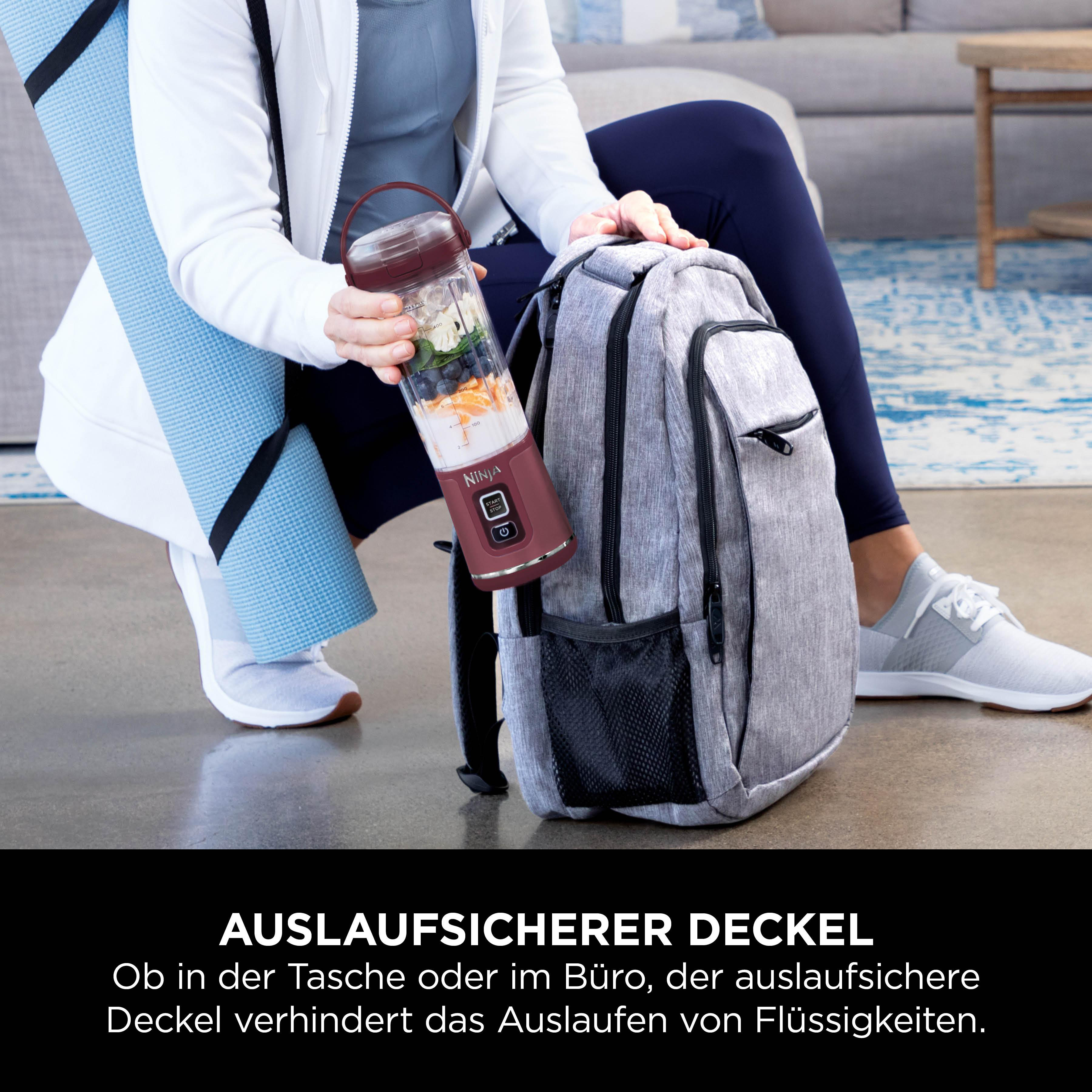 Person hält einen weinroten Mixer mit Zutaten neben einem Rucksack und einer Yogamatte. Der Mixer ist mit „NINJA“ beschriftet.