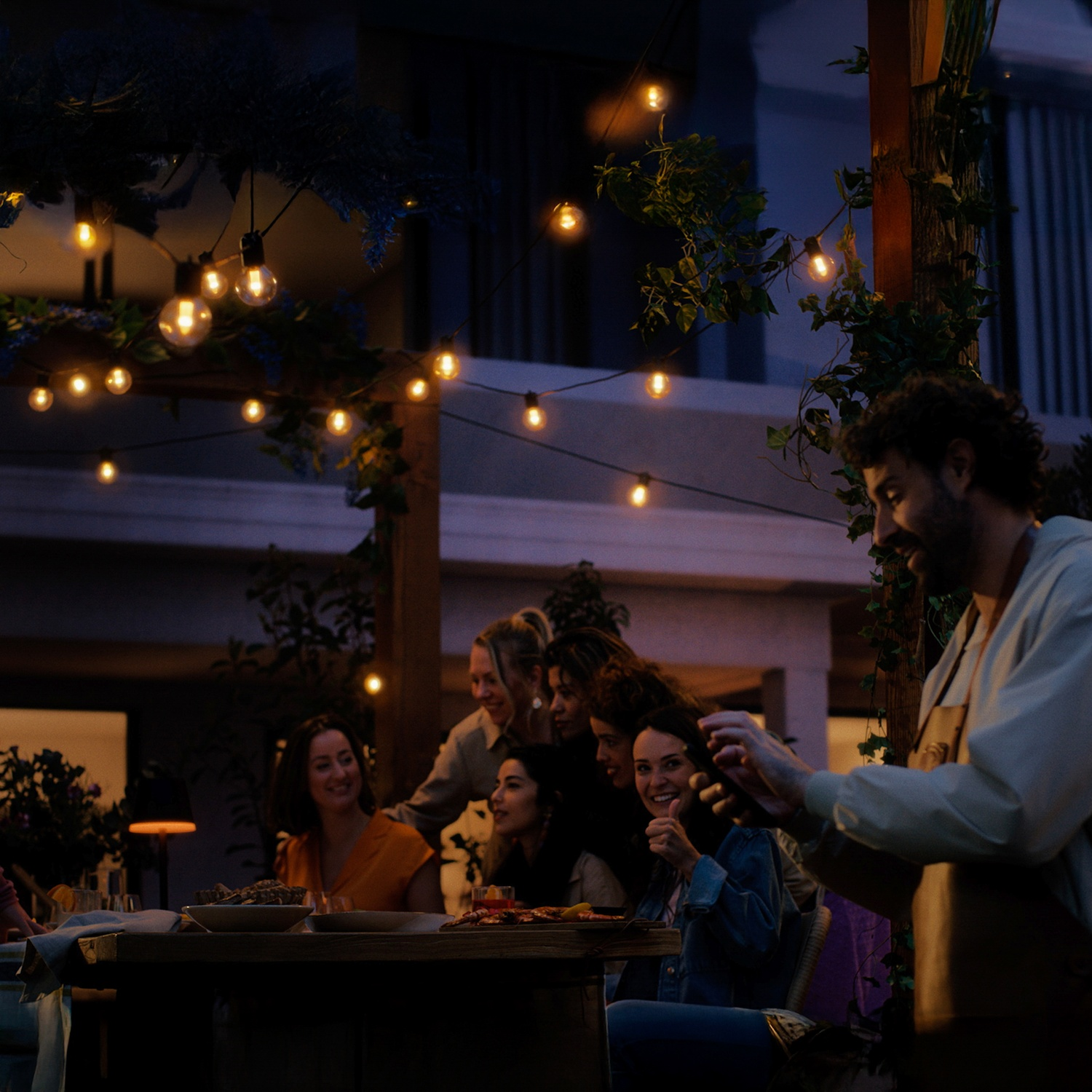 Group of people at a dinner party outdoors at night. String lights illuminate the scene. People are smiling and enjoying themselves.