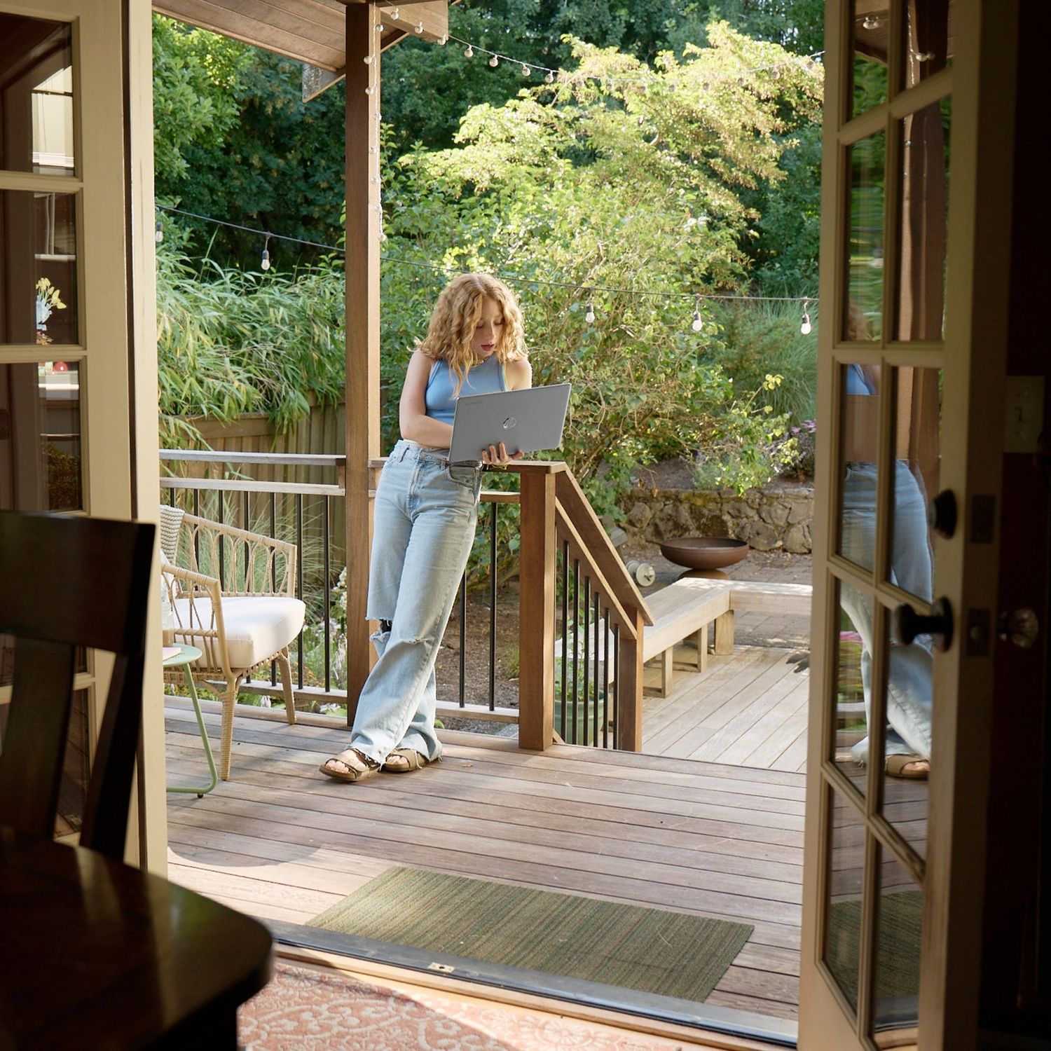 Woman on a porch with a laptop, looking at the screen. Wooden deck with a bench and greenery in the background.