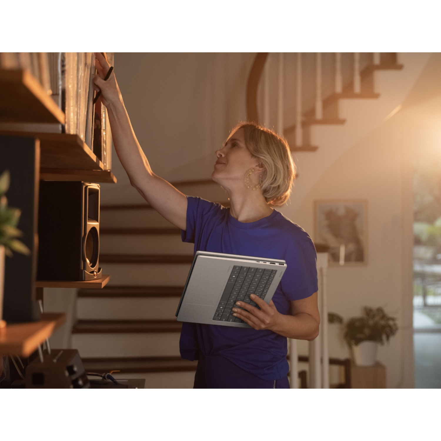 A woman in blue holds a laptop and reaches for a book on a shelf. Sunlight streams in, illuminating a staircase in the background.