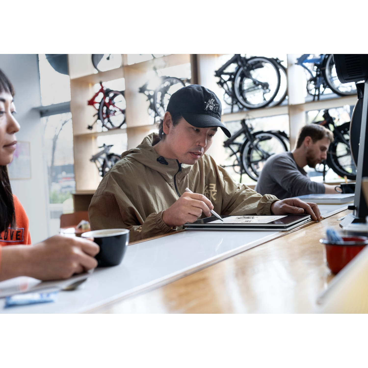 A person in a cap draws on a tablet. A woman holds a cup, and a man works at a computer. Bikes hang in the background.