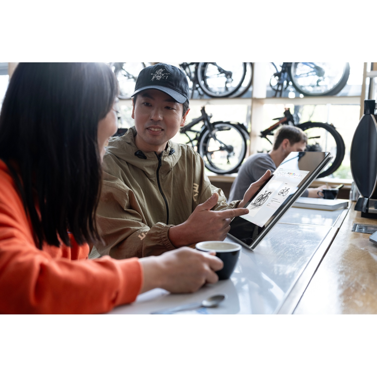 Two people looking at a tablet showing a bicycle. Man pointing, holding coffee. Bikes in background. Woman in orange.