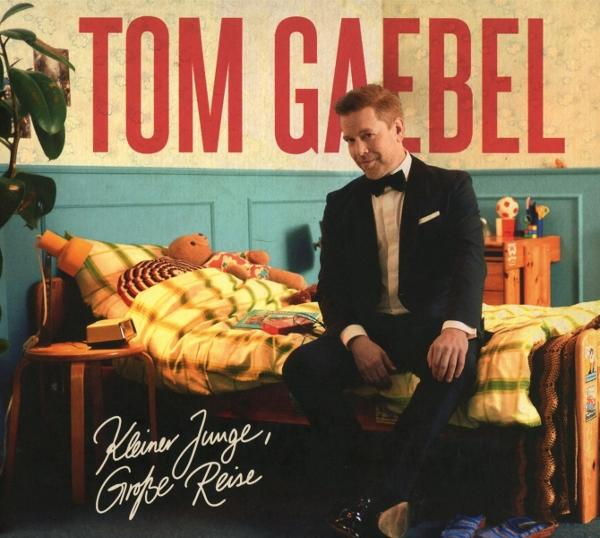 Tom Gaebel Albumcover. Mann im Anzug sitzt auf einem Bett mit Spielzeug und Teddybär. Roter Text 'TOM GAEBEL'. Albumtitel unten.